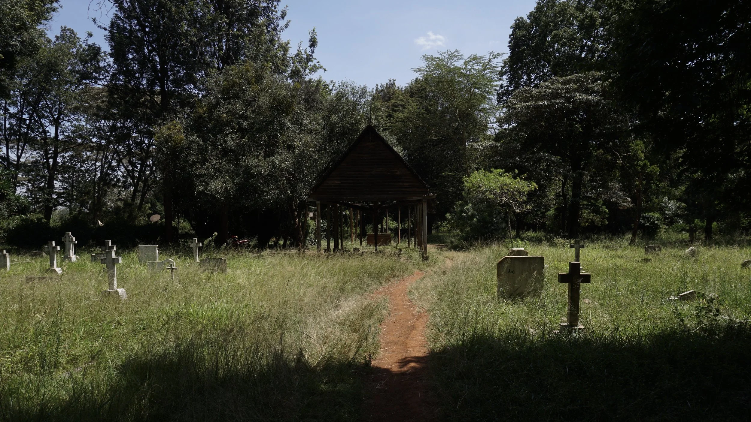 A small graveyard with white crosses and headstones, a dirt path leading to a wooden shelter, surrounded by tall grass and trees.