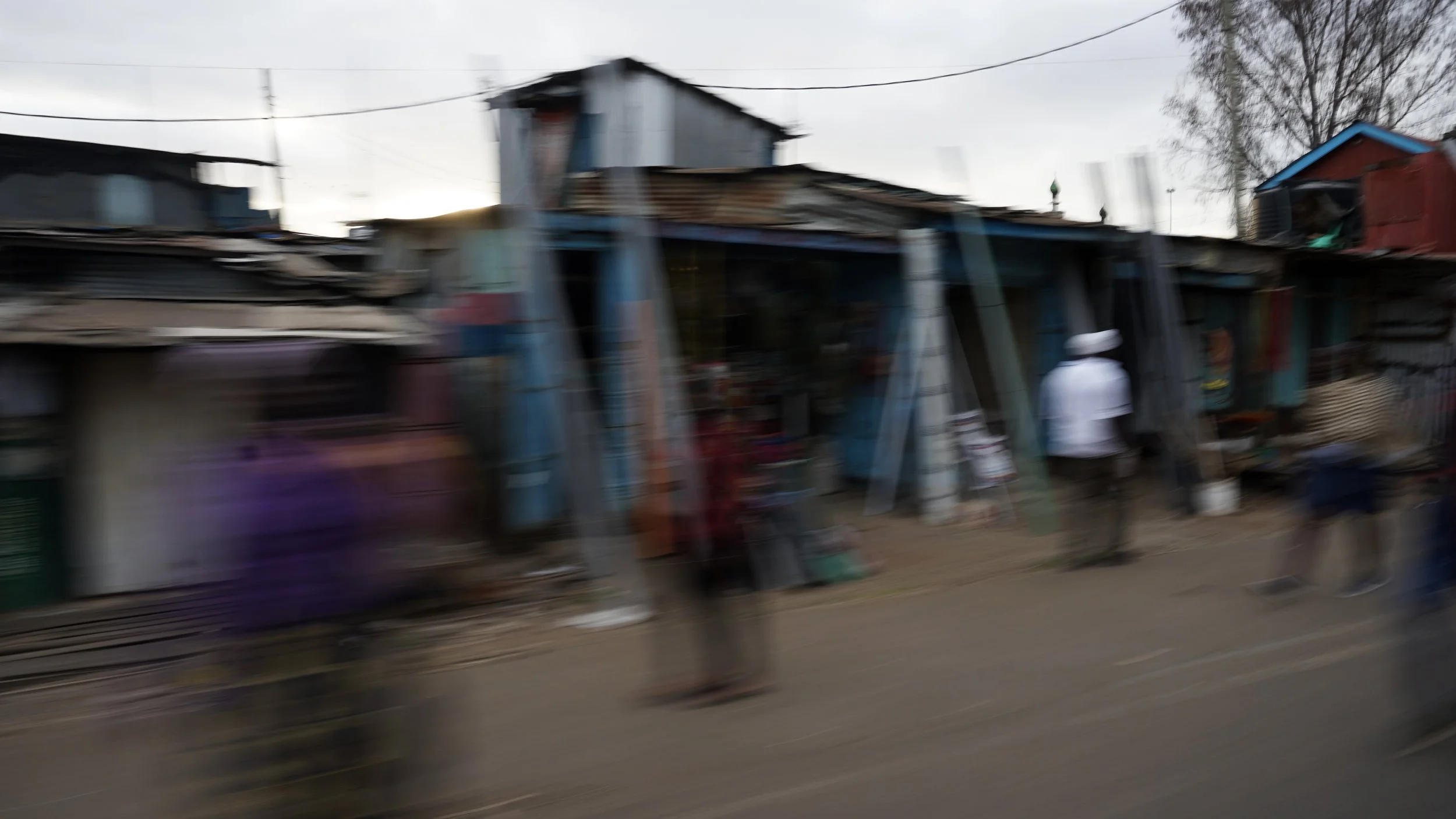 Blurred image of a street scene with small shops and people walking, with overcast sky and some trees in the background.