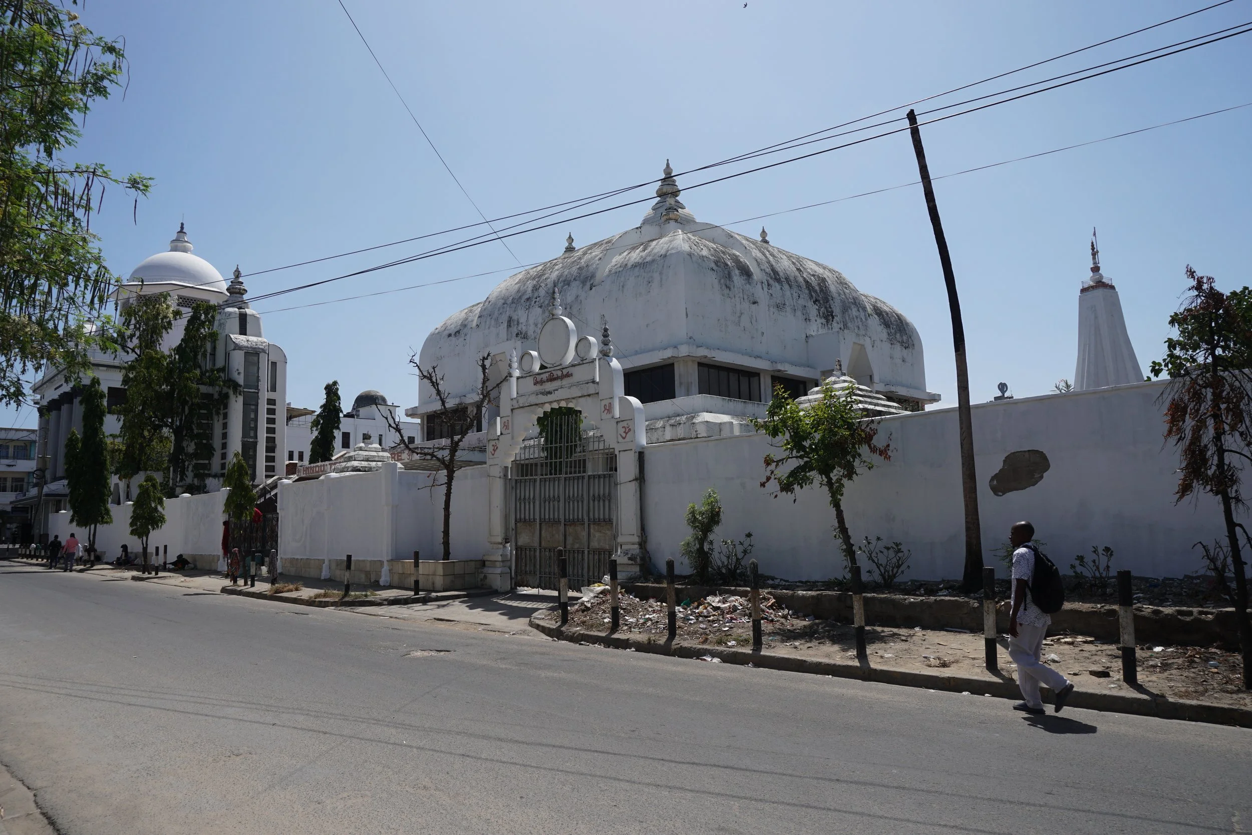 A large white religious building with dome-shaped roof, surrounded by a white wall and gate, situated along a paved street with trees and pedestrians, under a clear blue sky.