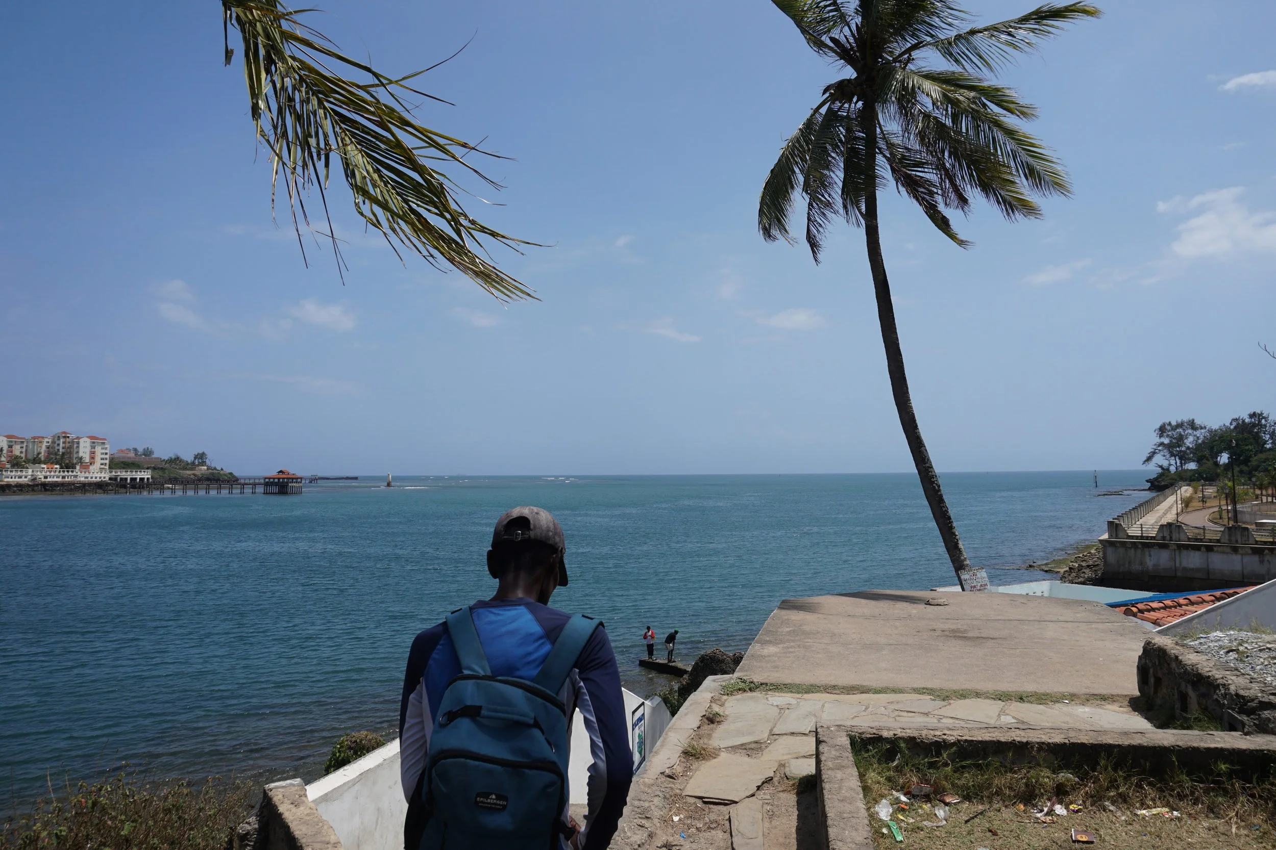 A person with a backpack and cap stands on a pathway overlooking a blue ocean, with palm trees and a few people near the shore. There are buildings in the distance under a partly cloudy sky.