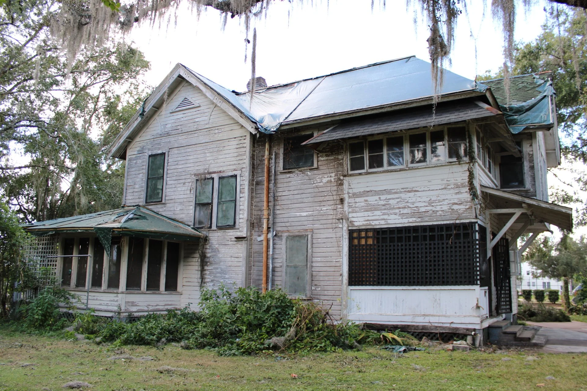 A run-down, old, two-story white wooden house with peeling paint, broken windows, and a sagging roof with blue tarp coverings. The house is surrounded by overgrown plants and trees with hanging moss.