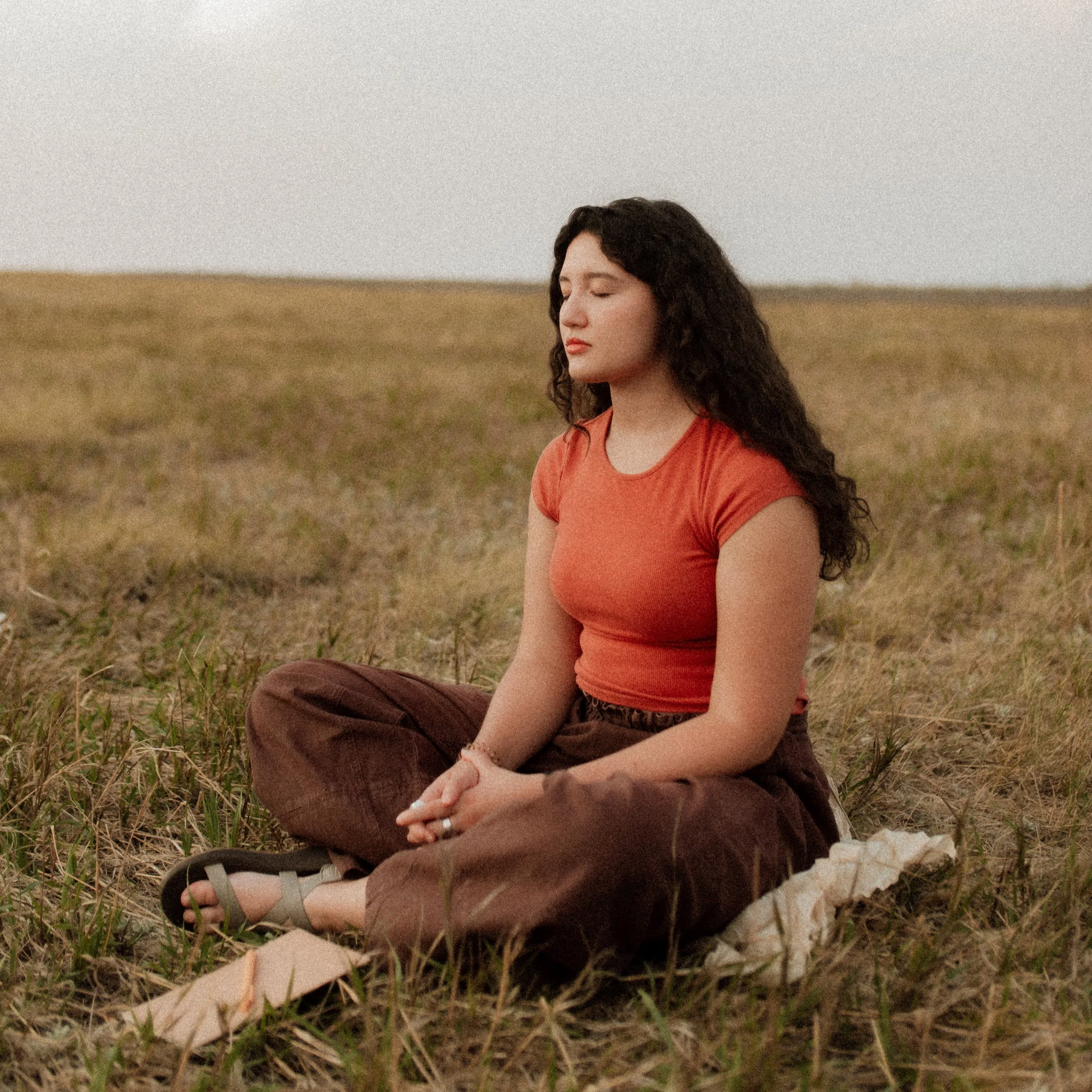 A young woman with curly dark hair, wearing an orange shirt and brown pants, sitting on the ground outdoors with closed eyes, meditating in a field