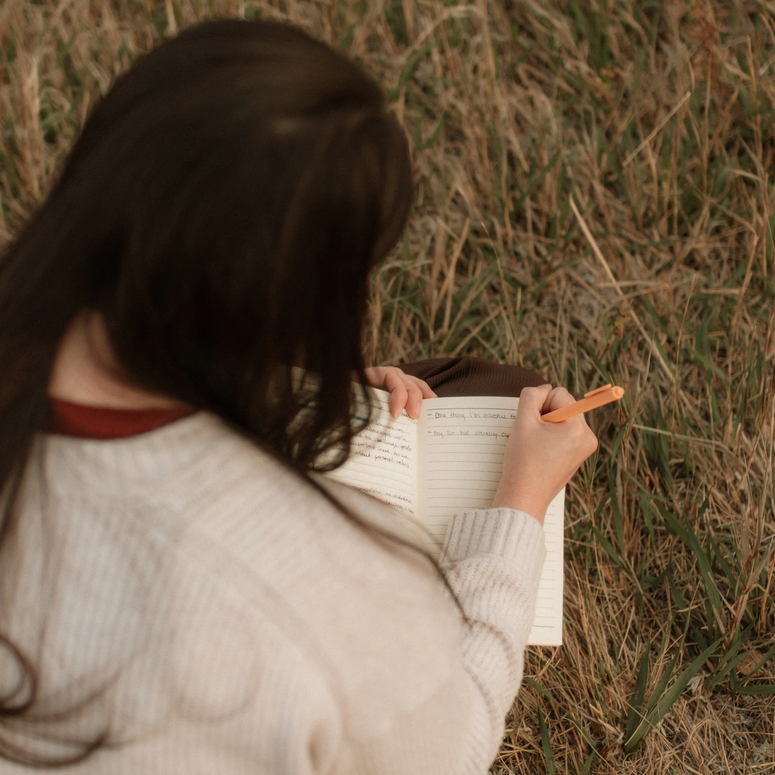 A person sitting outdoors on grass, writing in a journal with a pen.