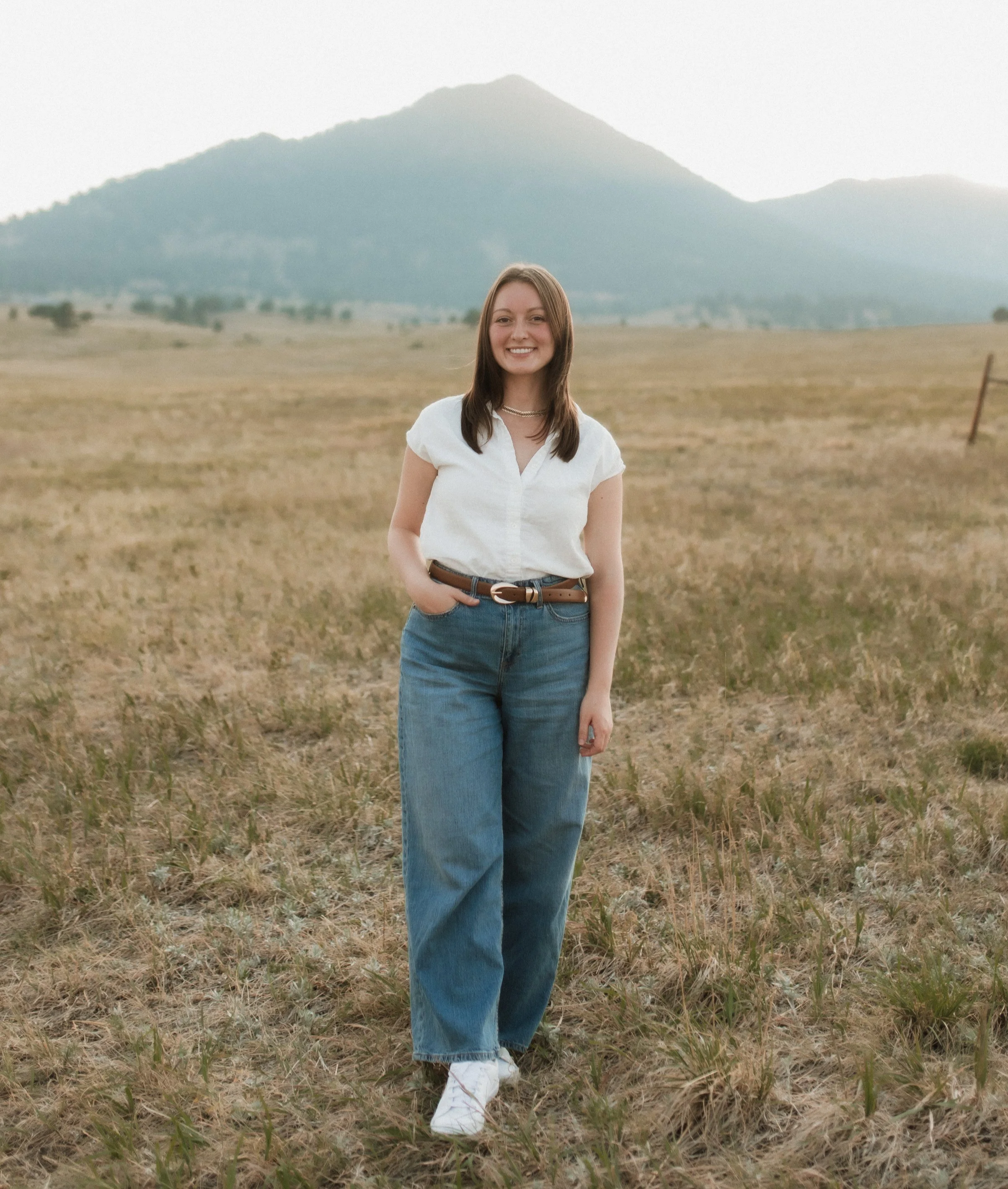 Young woman smiling outdoors in a field with tall grass, wearing a white shirt, blue jeans, and white shoes, with a mountain in the background.