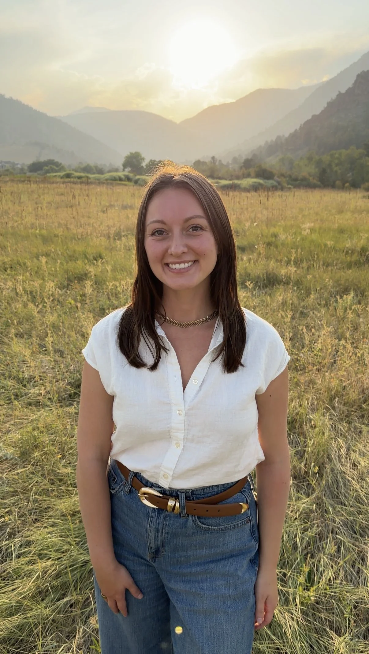 A woman with brown hair and a white shirt standing in a grassy field at sunset with mountains in the background.