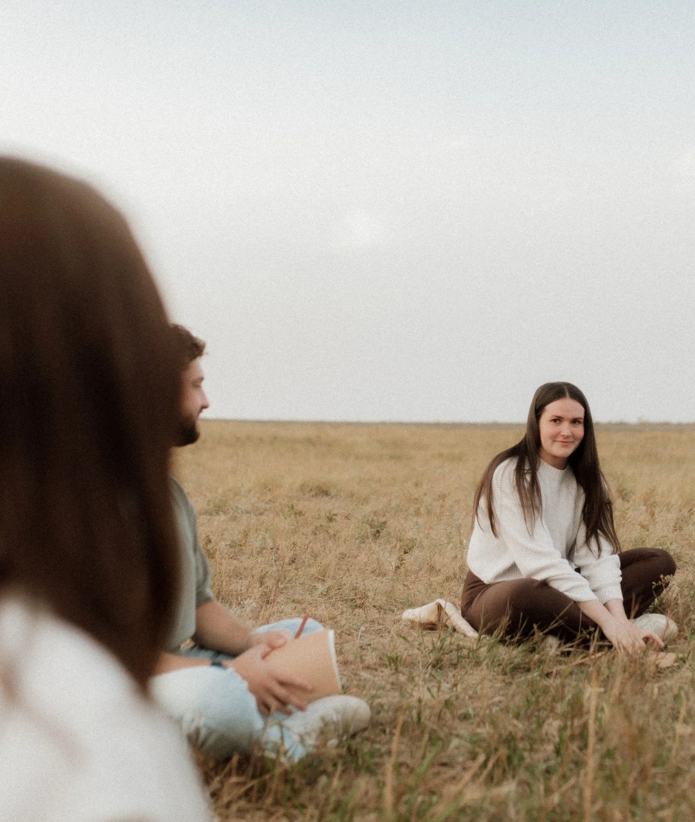 Two women sitting on a grassy field during daytime, one with long dark hair and white sweater, the other with long dark hair and green shirt, under an overcast sky.