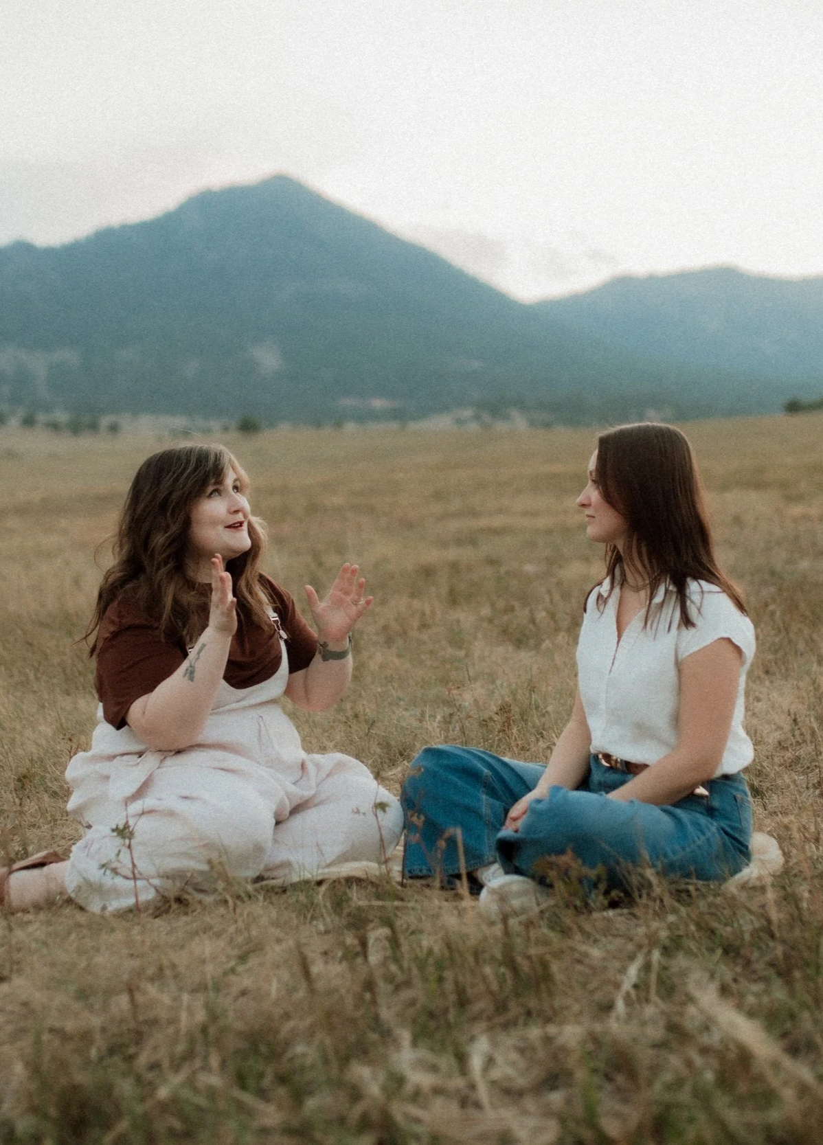 Two women sitting on the grass in a field, engaging in a conversation, with mountains in the background.