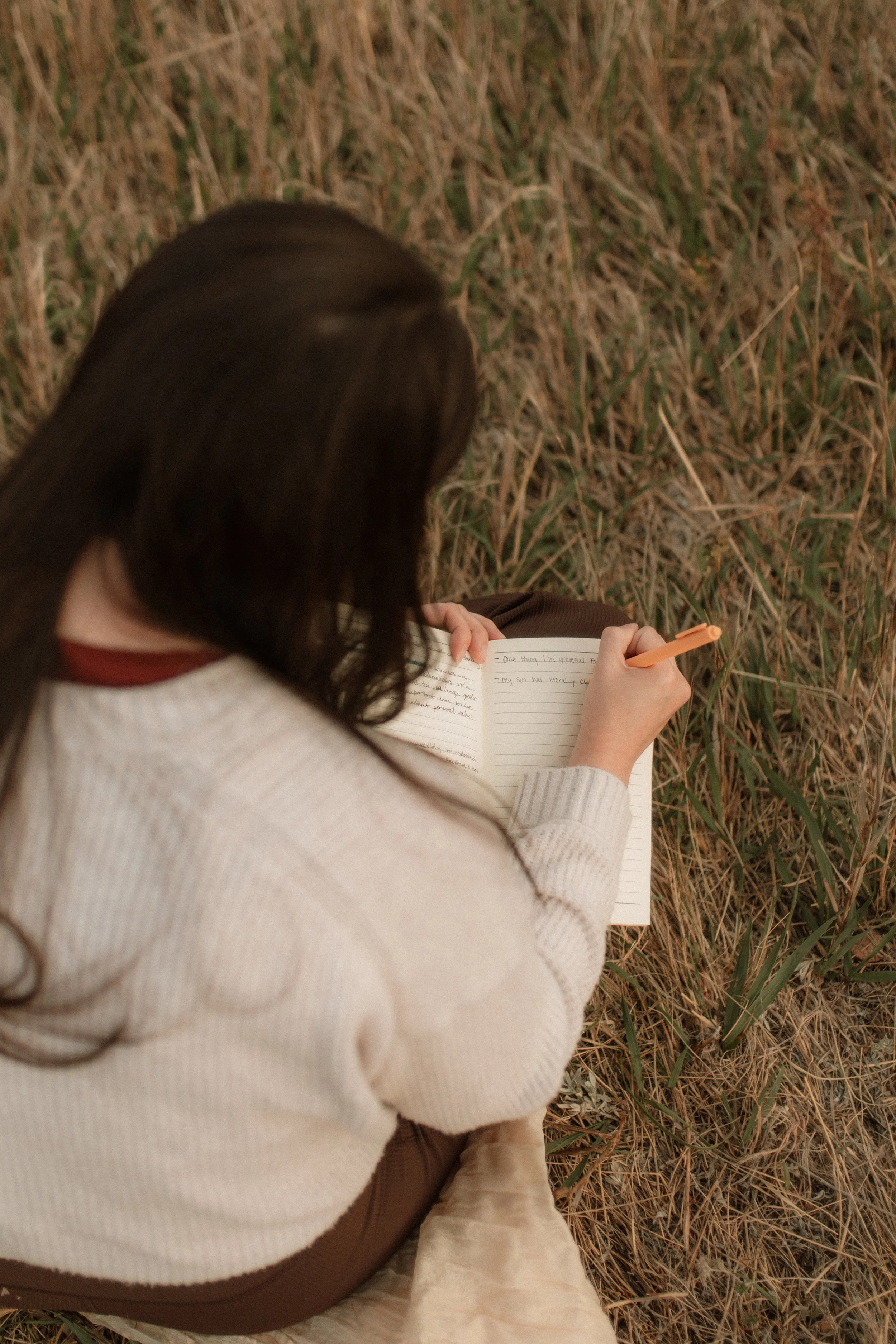 Person with dark hair sitting on grass, writing in a journal with an orange pen, wearing a white sweater.