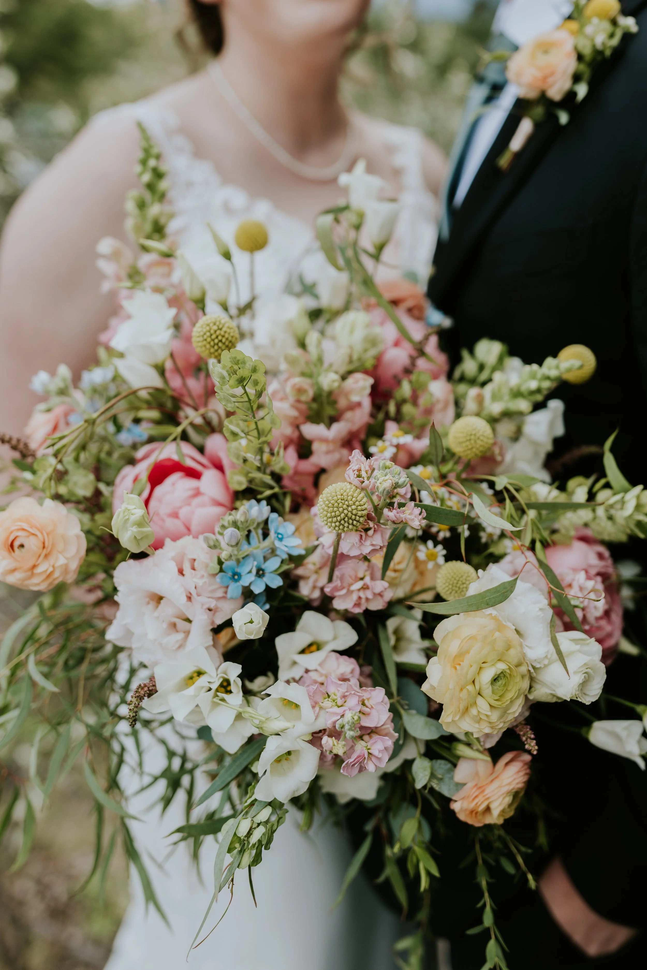 Close-up of a wedding bouquet with pink, white, and yellow flowers, with a bride and groom partially visible in the background.