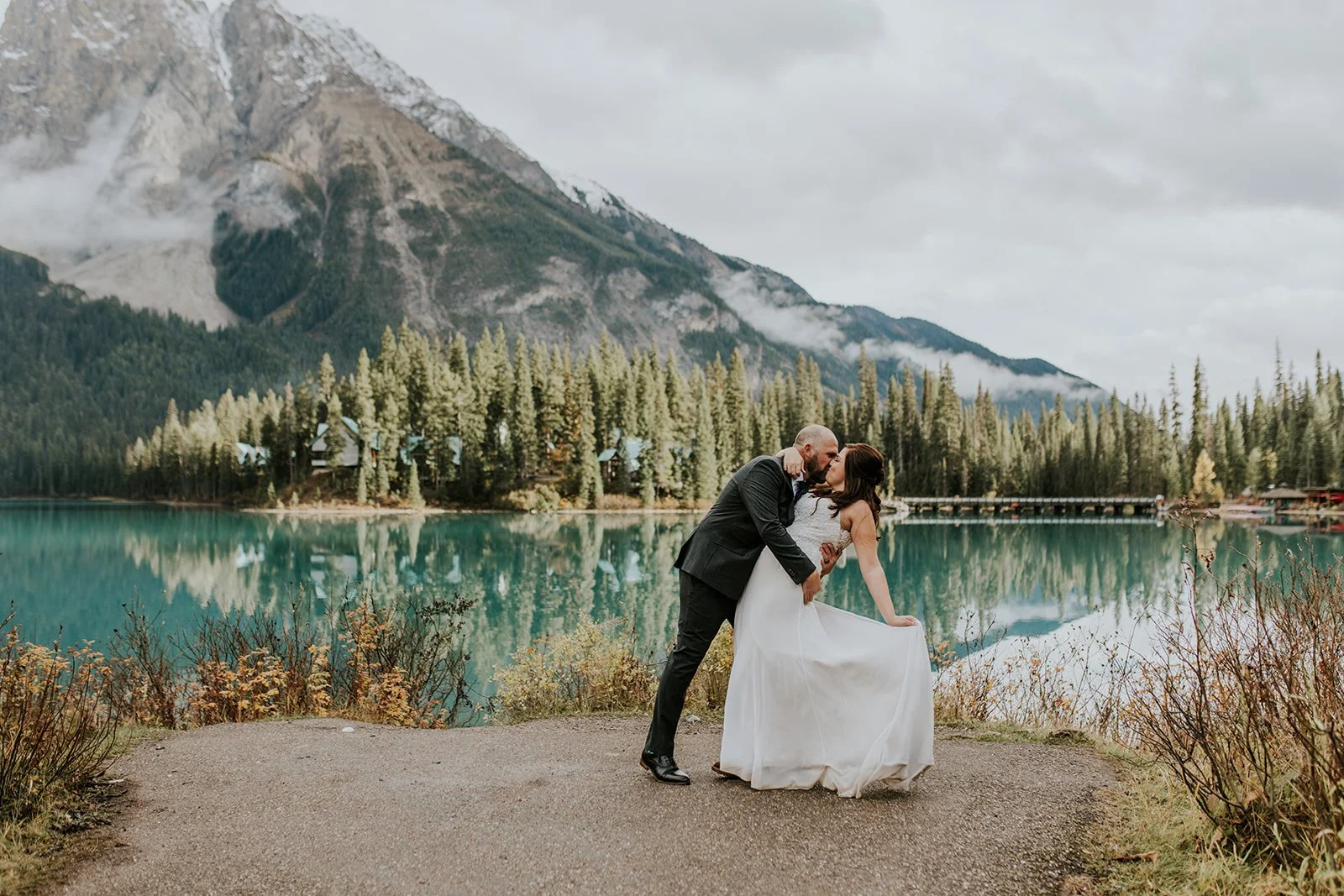 A couple dressed in formal wedding attire sharing a kiss by a lake with mountains and evergreen trees in the background.