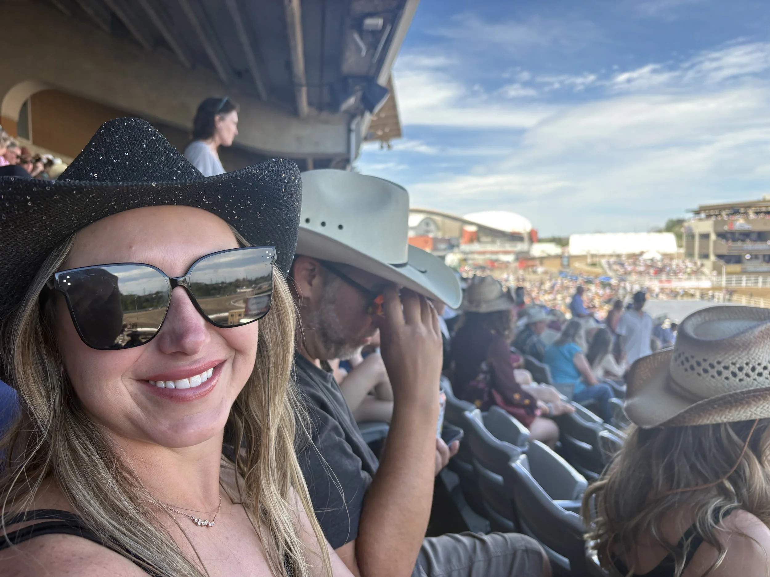 A woman with long blonde hair, wearing a black hat, sunglasses, and a necklace, smiling as she takes a selfie on a boat. There are other people sitting behind her, some with hats and sunglasses, and the sky is partly cloudy.