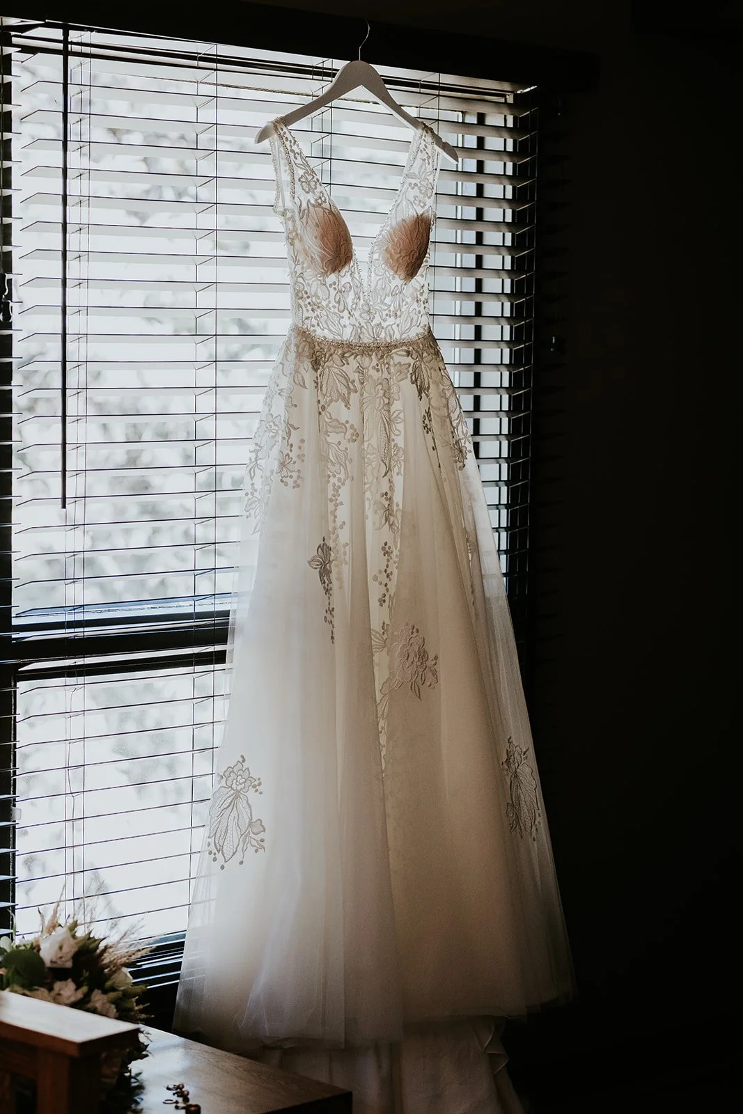 A white wedding dress with lace floral details hanging on a hanger in front of a window with blinds, backlit by natural light.