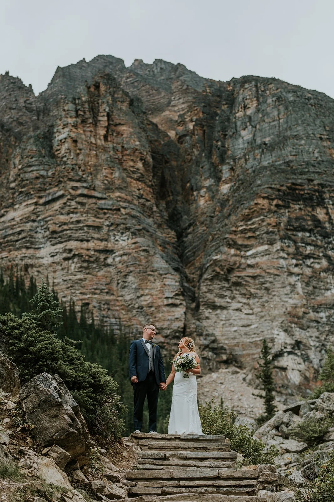 A bride and groom holding hands and looking at each other in a mountain setting, with steep rocky cliffs and evergreen trees in the background.