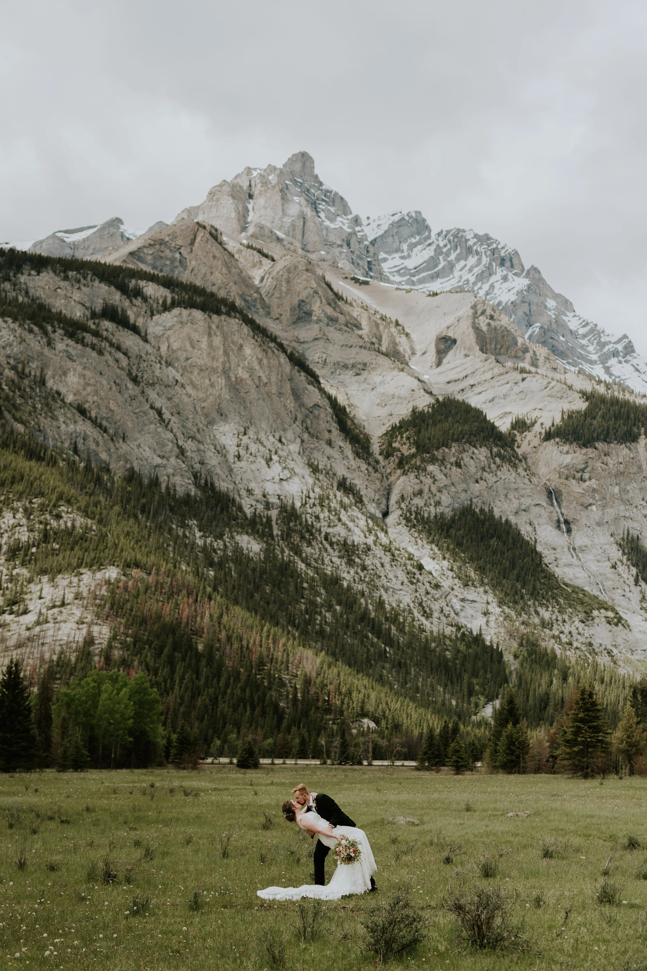 A bride and groom kissing on a grassy field with mountains in the background.