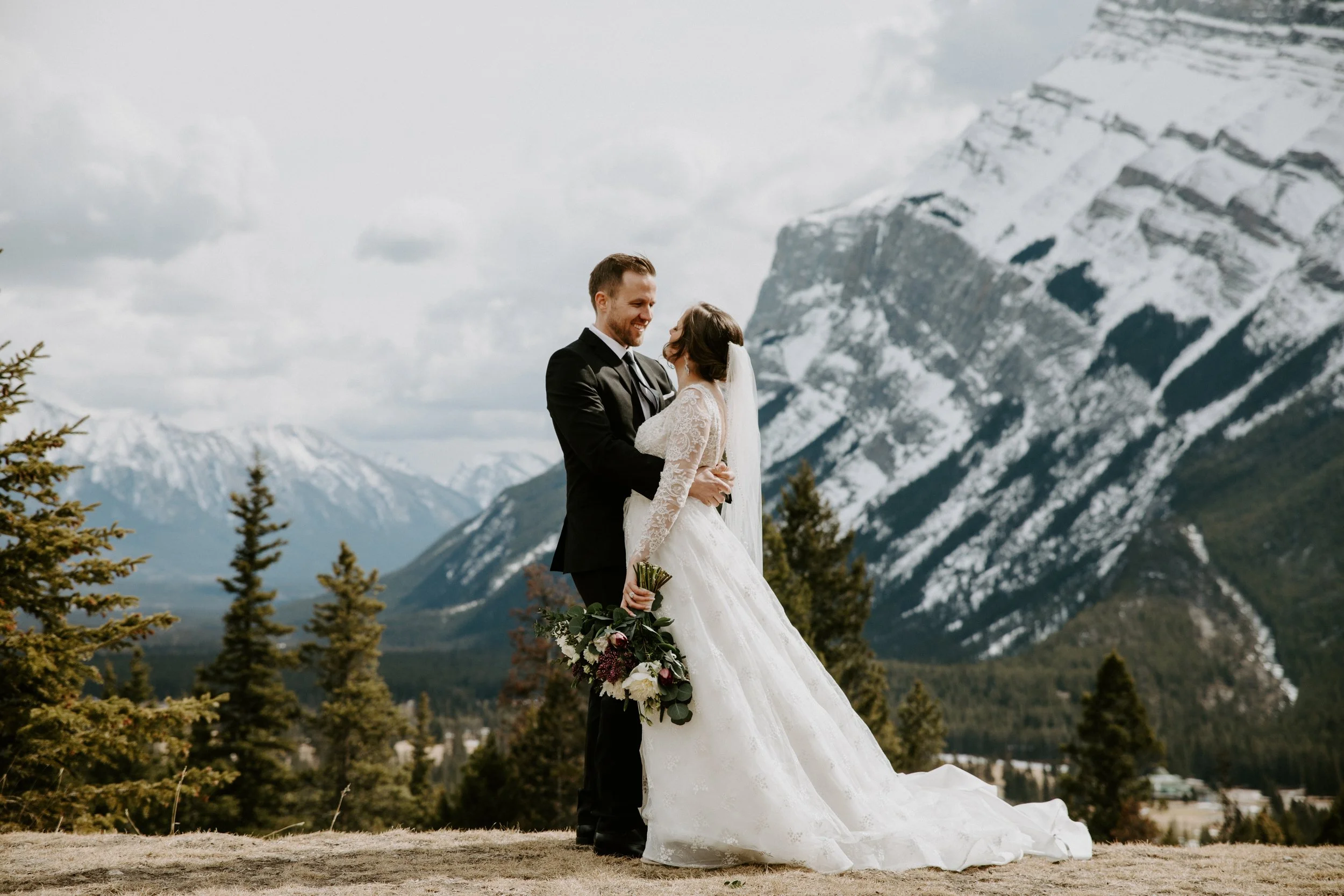 A bride and groom standing outdoors in front of a mountainous landscape, smiling and gazing at each other on their wedding day. The bride is wearing a white lace wedding dress and veil, holding a bouquet, and the groom is in a black tuxedo.