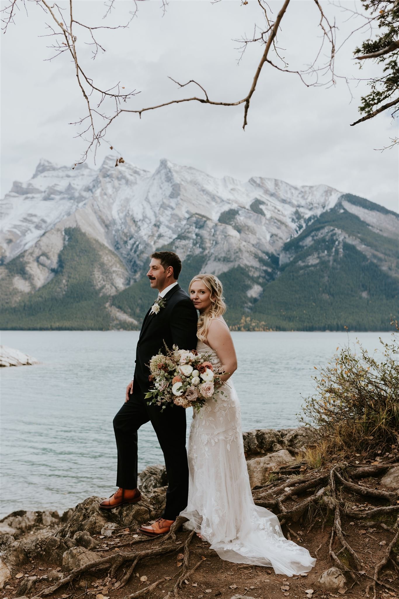 A couple in wedding attire standing on rocks by a lake with mountains and cloudy sky in the background. The groom wears a black suit and brown shoes, while the bride wears a white lace dress and holds a bouquet of flowers.