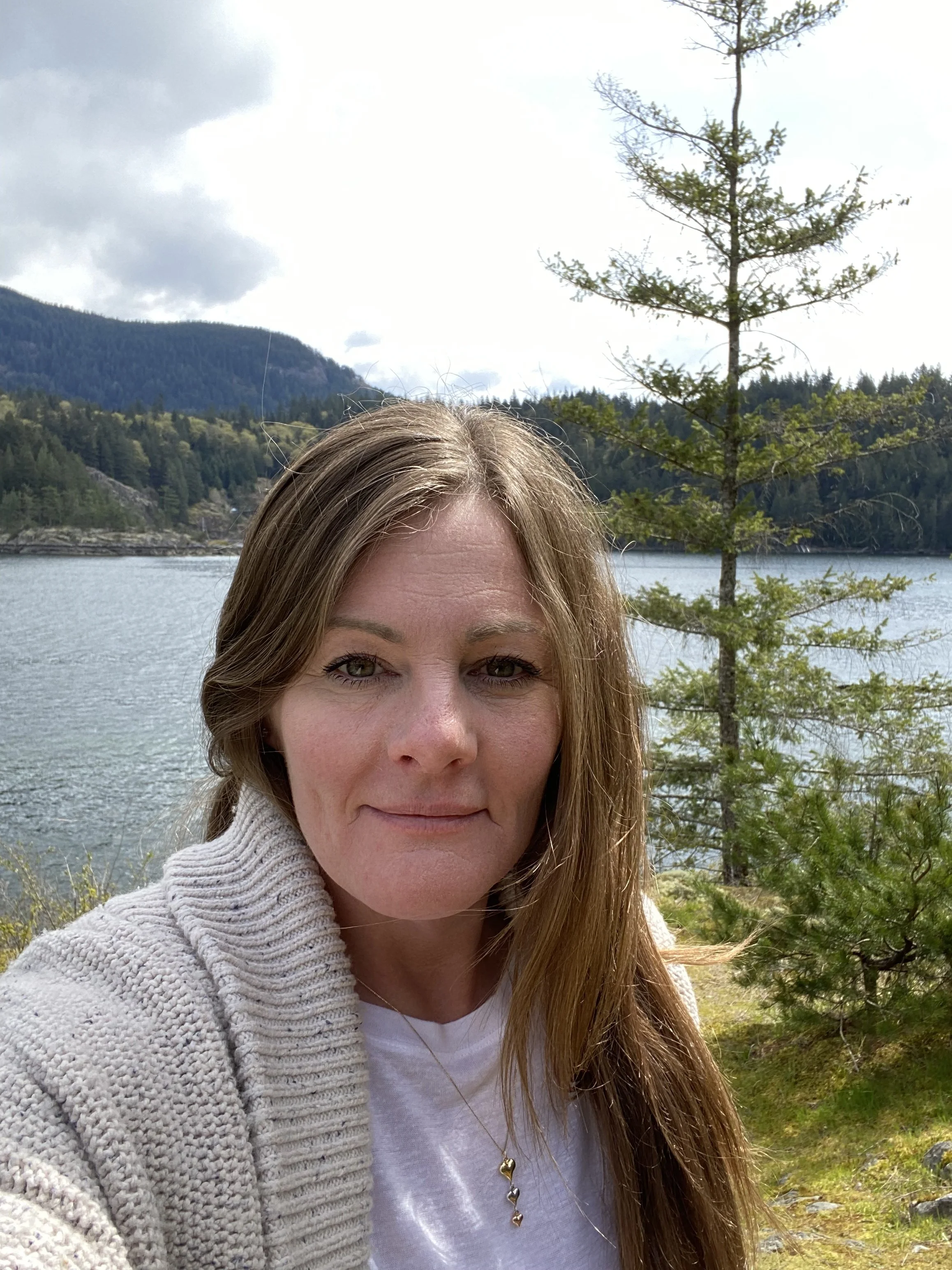 A woman with long light brown hair taking a selfie outdoors, with a lake, evergreen trees, and mountains in the background under a partly cloudy sky.
