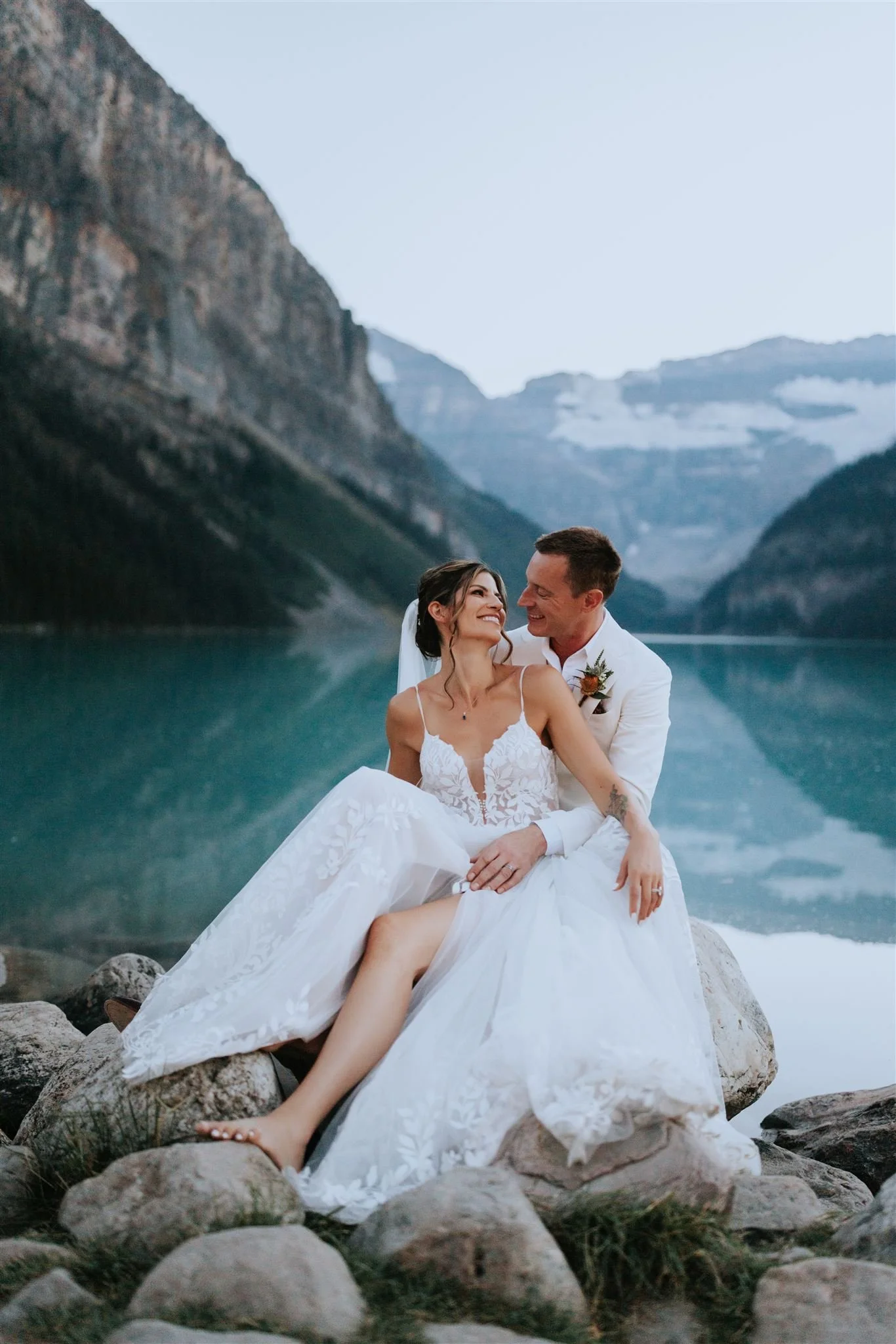 A bride and groom sitting on rocks by a lake surrounded by mountains, with the bride wearing a white wedding dress and the groom in a white shirt. They are smiling and looking at each other with happiness.