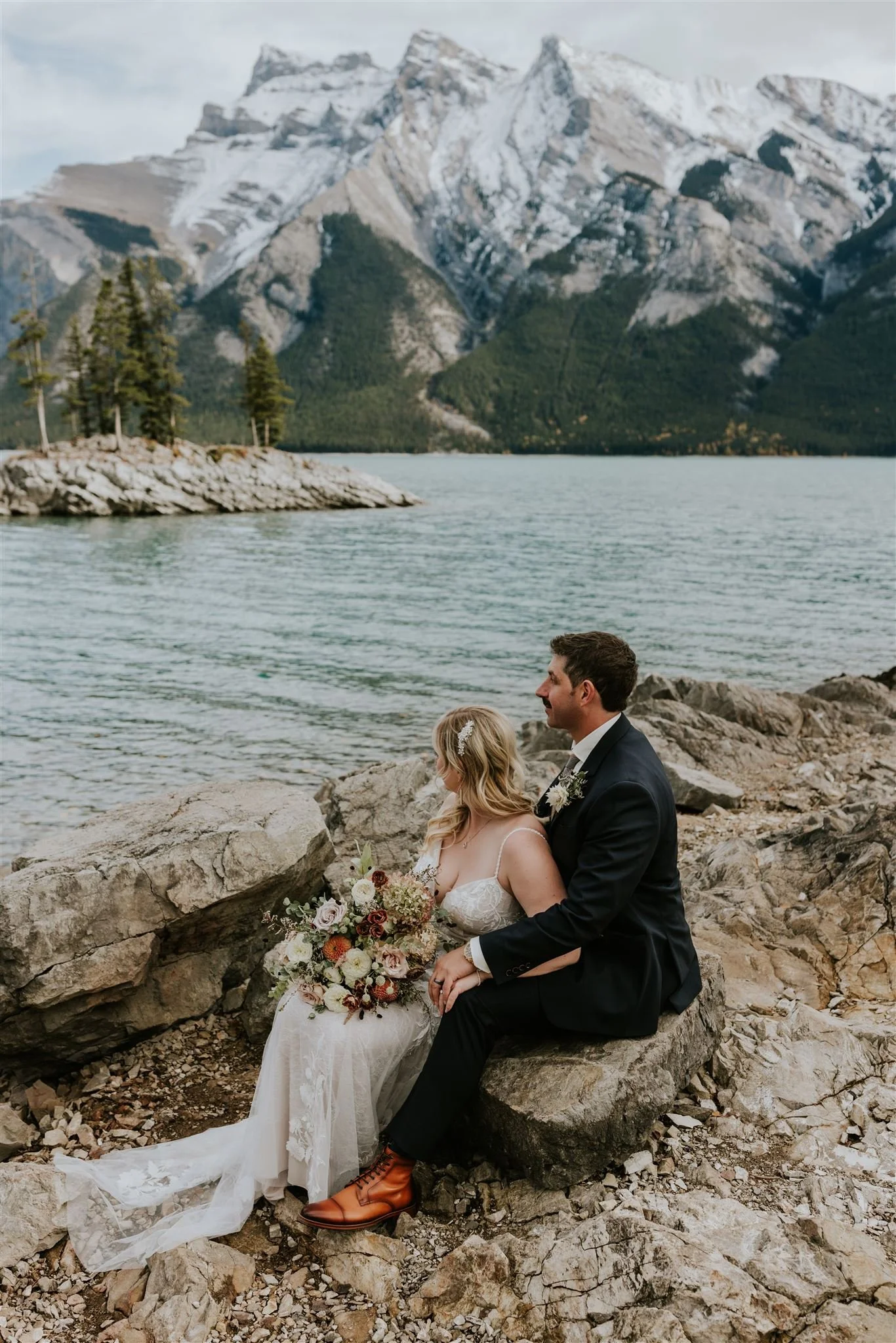 A bride and groom sitting on rocks by a lake, surrounded by mountains and trees, during their wedding ceremony.