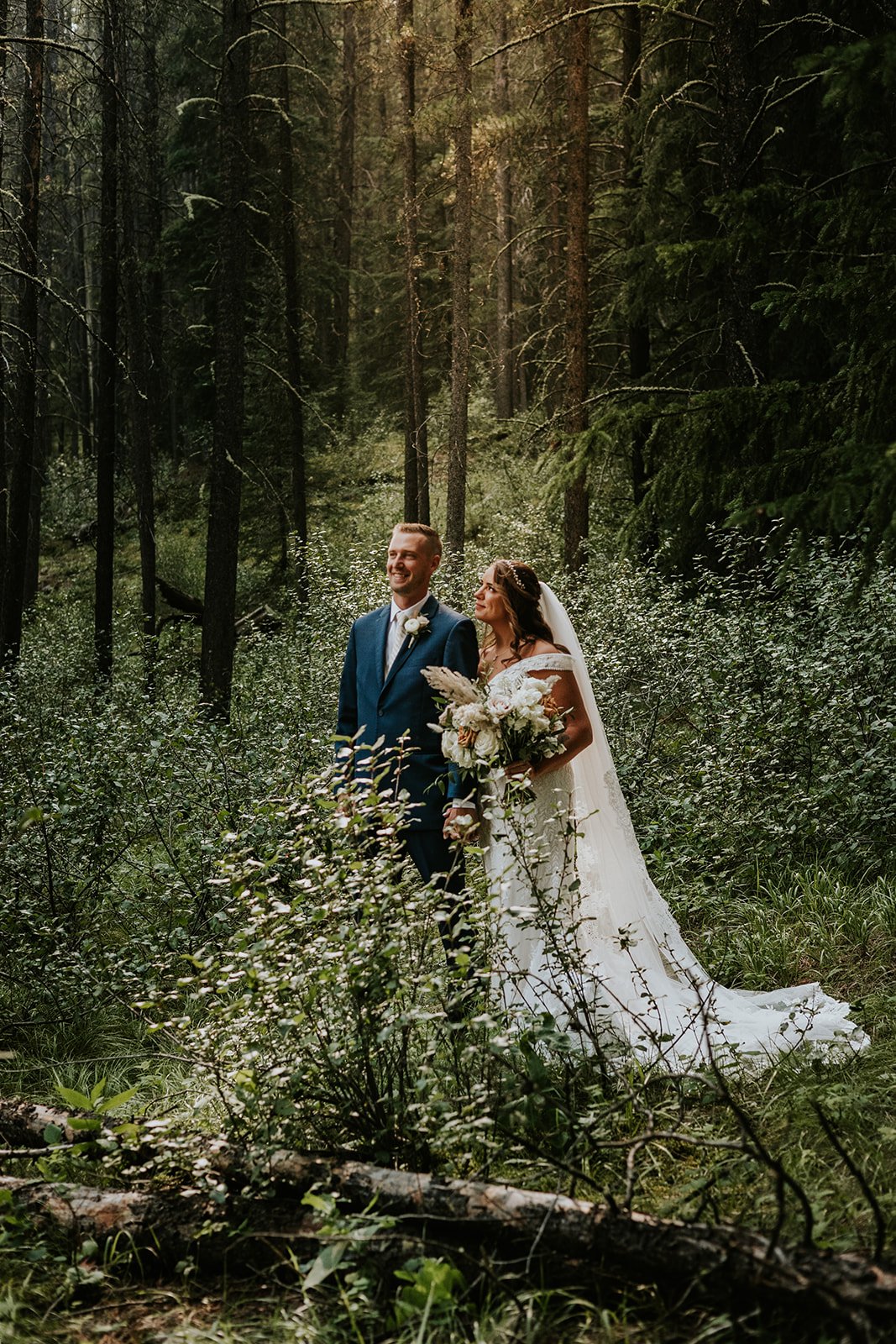 A bride and groom standing hand in hand in a forest, with the bride holding a bouquet of white flowers and greenery, sunlight filtering through trees.