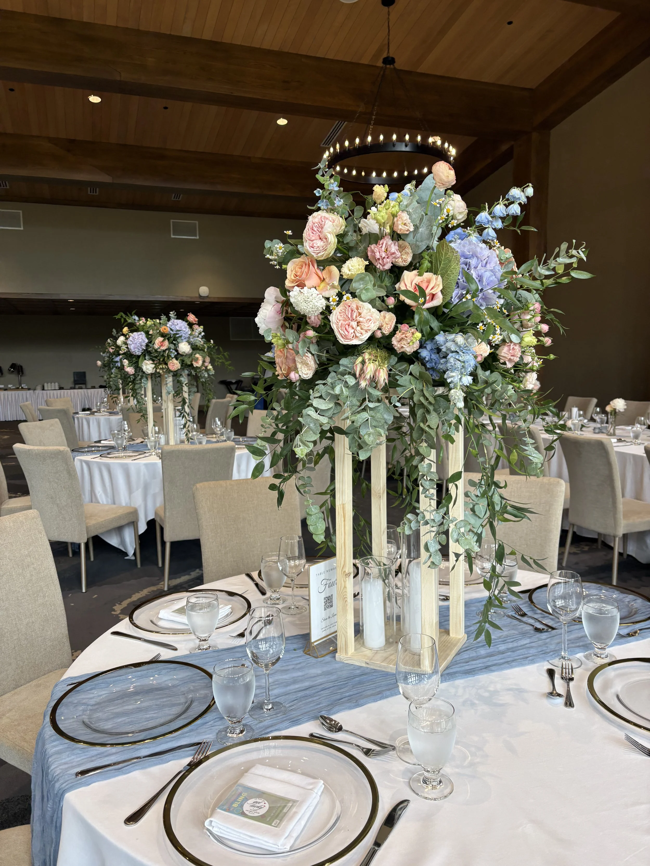 Elegant banquet table setting with white tablecloth, floral centerpiece, glassware, white napkin, and black-edged plates, in a decorated event space.