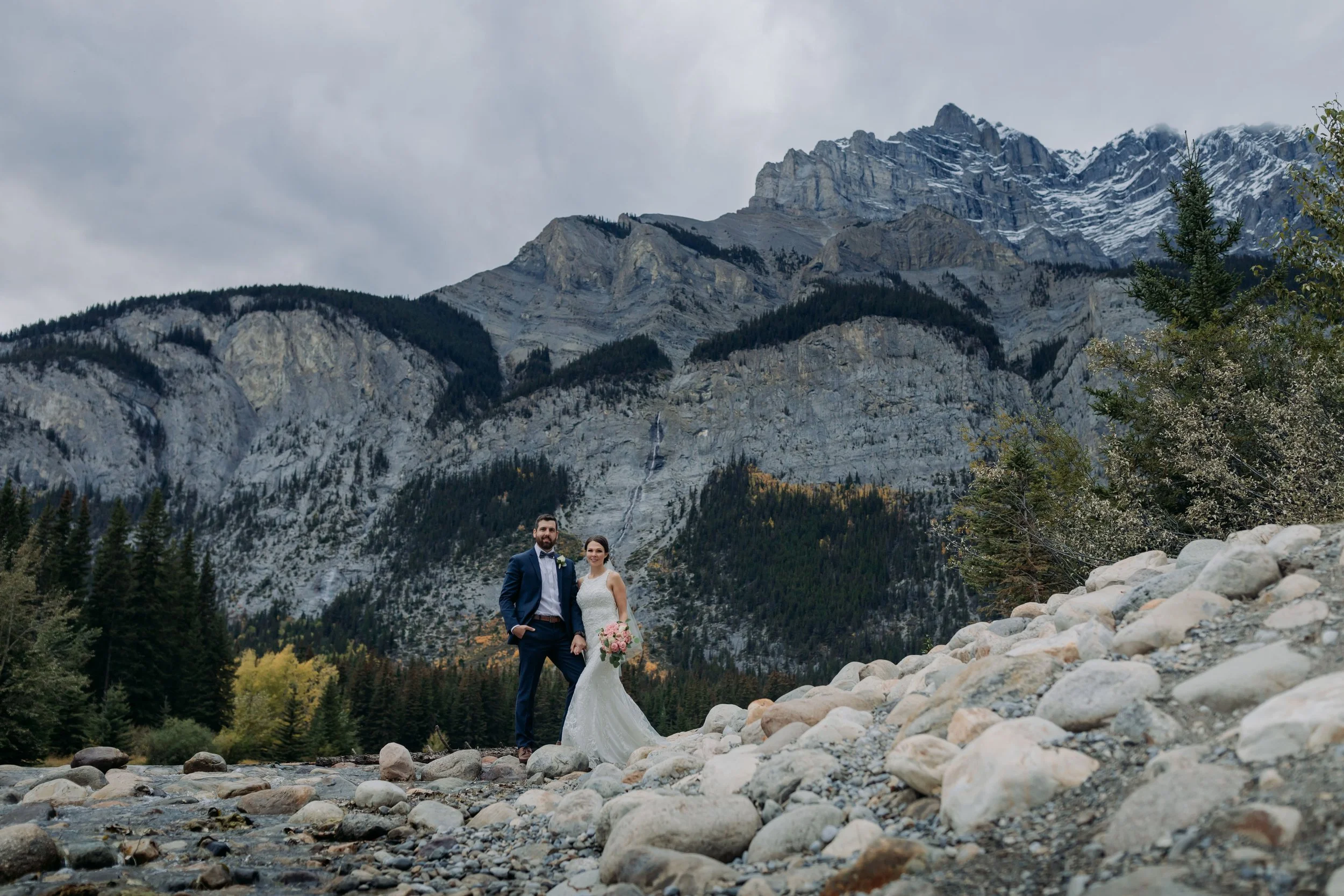 A bride and groom in wedding attire standing on river rocks in a mountainous landscape with forested hills and snow-capped peaks in the background.