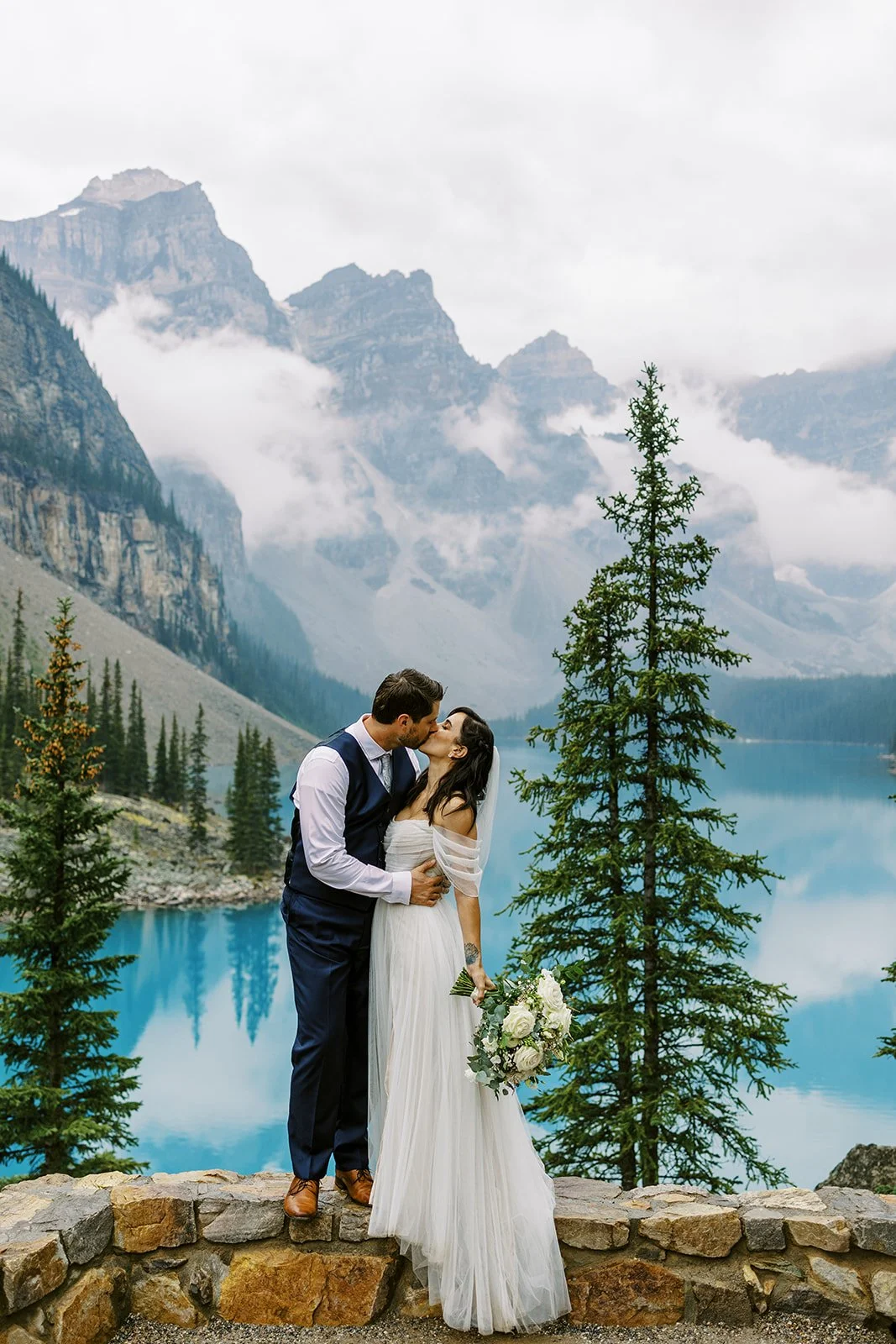 A couple in wedding attire sharing a kiss by a mountain lake with pine trees and mountains in the background.
