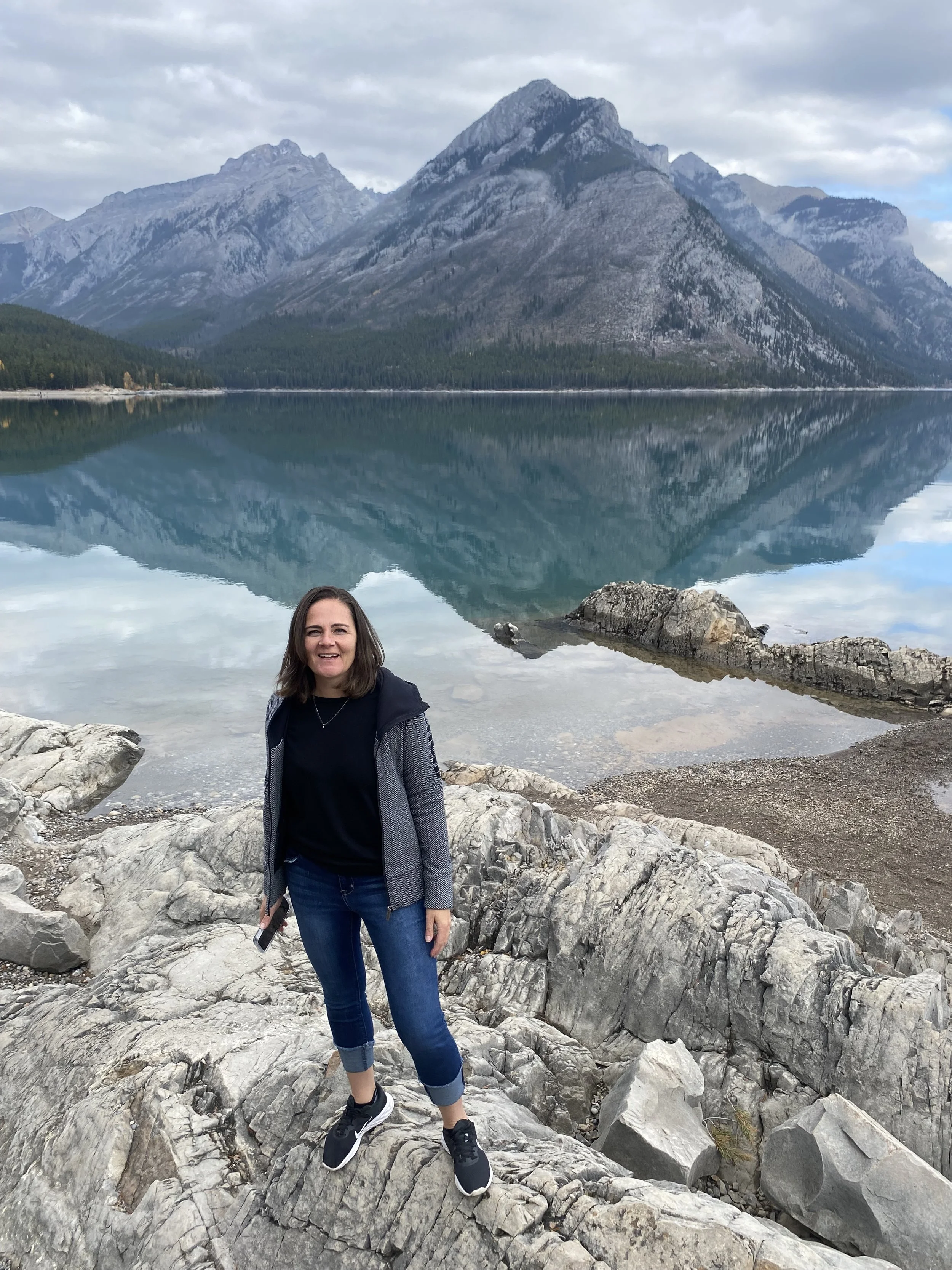 A woman standing on large rocks by a lake with calm water, reflecting a mountain range with a tall mountain and cloudy sky in the background.