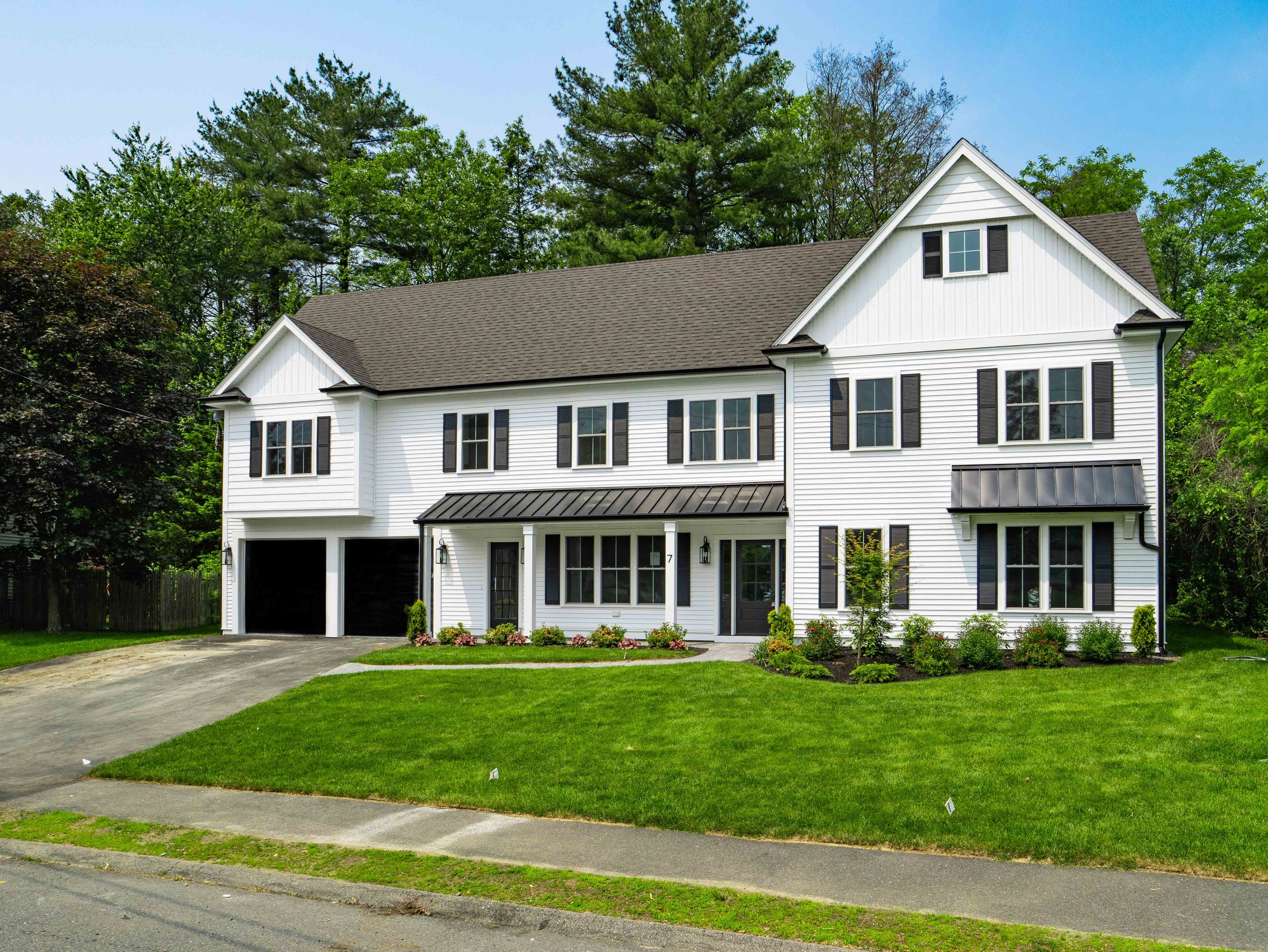 A two-story white house with black shutters and a brown roof, surrounded by green trees and a neatly manicured lawn.