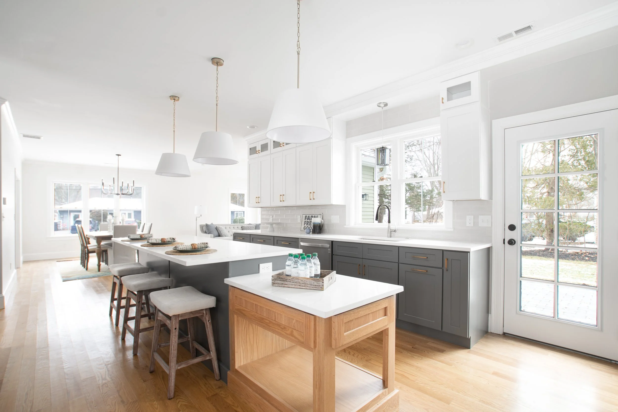 Bright kitchen with white and gray cabinets, a white island, wooden stools, and hardwood floors, with dining and living areas in the background.