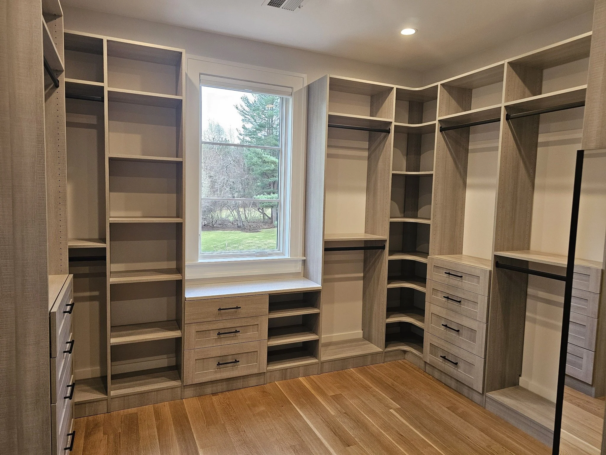 Empty walk-in closet with built-in cabinets, shelves, and hanging rods, a window showing trees outside, and hardwood flooring.