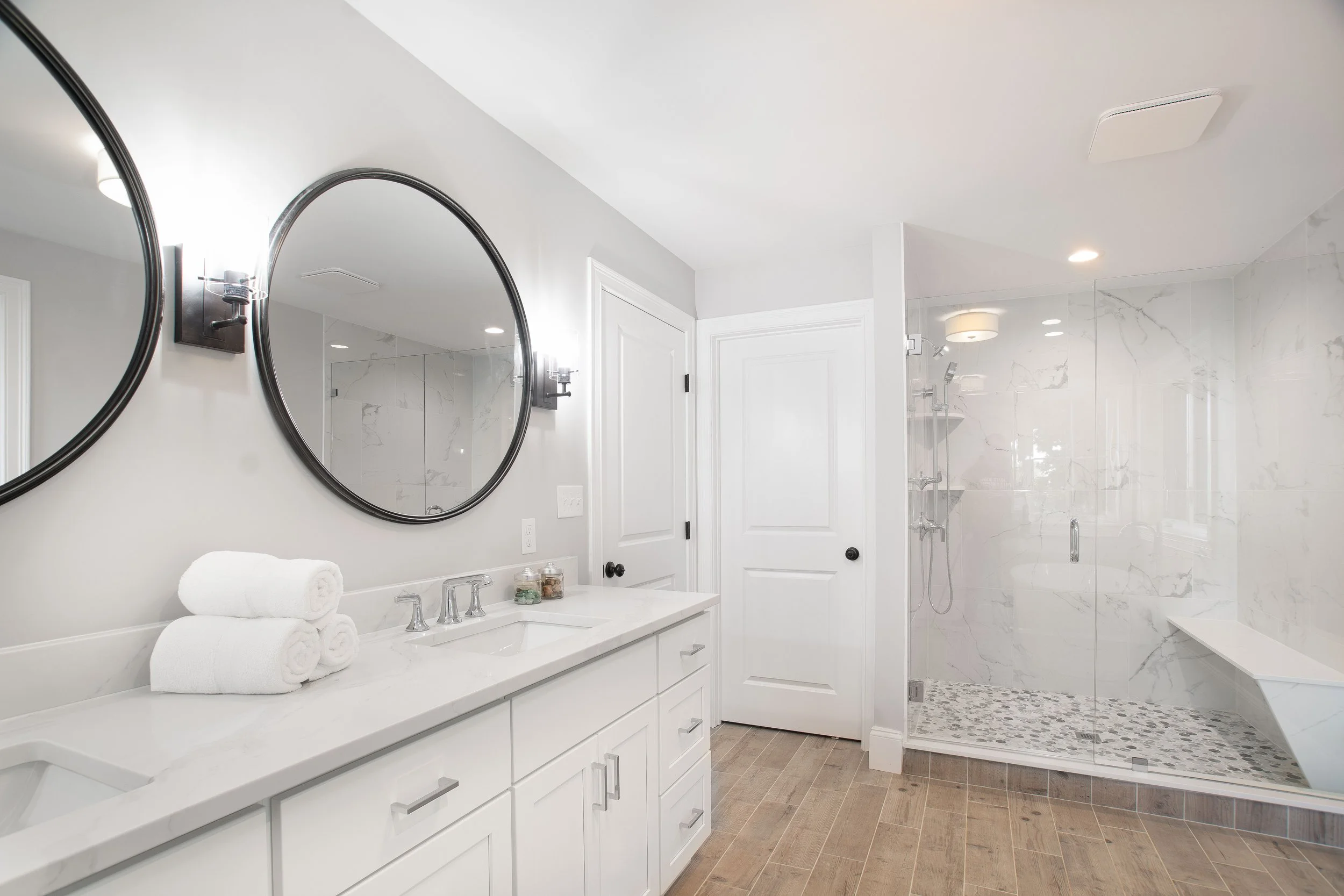 Modern bathroom with white double vanity, two round mirrors, and a glass-enclosed shower with marble tiles and wooden floor.