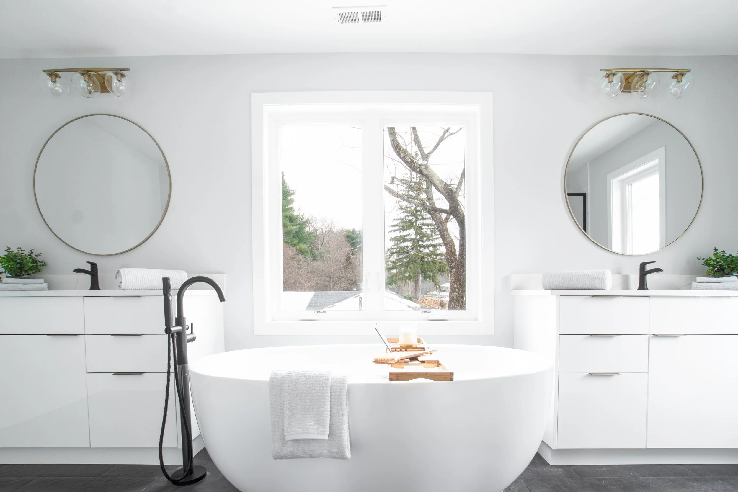 Bright, modern bathroom with a white oval bathtub in front of a large window. Two white vanities with black faucets, round mirrors, and matching decor on either side. Minimal decor with small plants and towels.