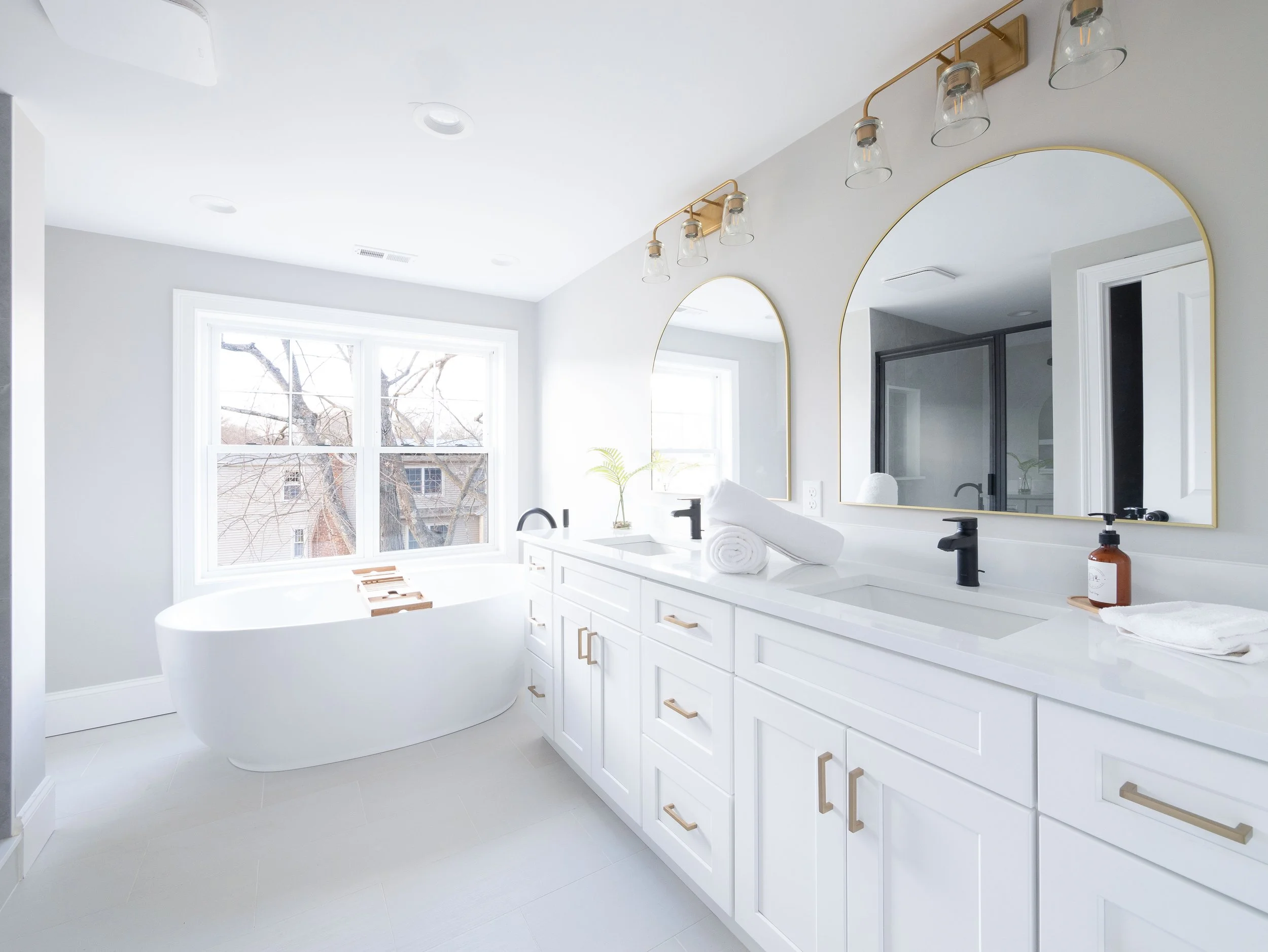 Bright modern bathroom with a white bathtub by a large window, white vanity with black faucets, oval mirrors with gold frames, and minimalist decor.