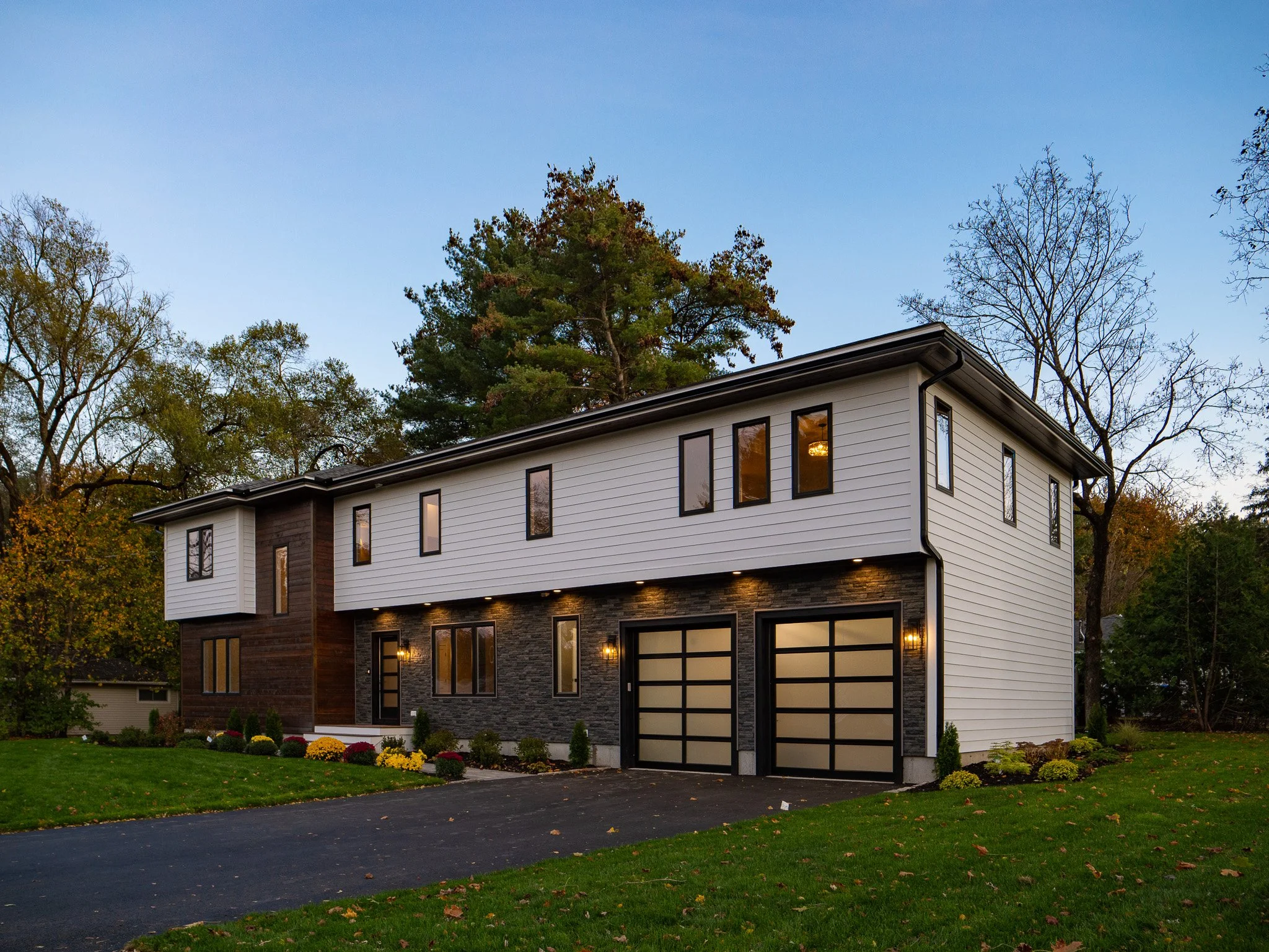 Modern two-story house with black and white exterior, two garage doors with frosted glass, surrounded by a well-maintained lawn and trees, during dusk.