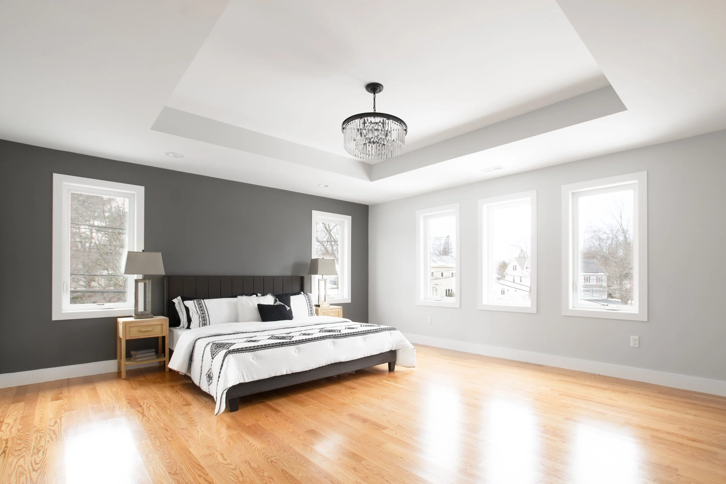 A modern bedroom with gray accent wall, white walls, wooden floors, a bed with black and white bedding, and two nightstands with lamps. Large windows provide natural light.