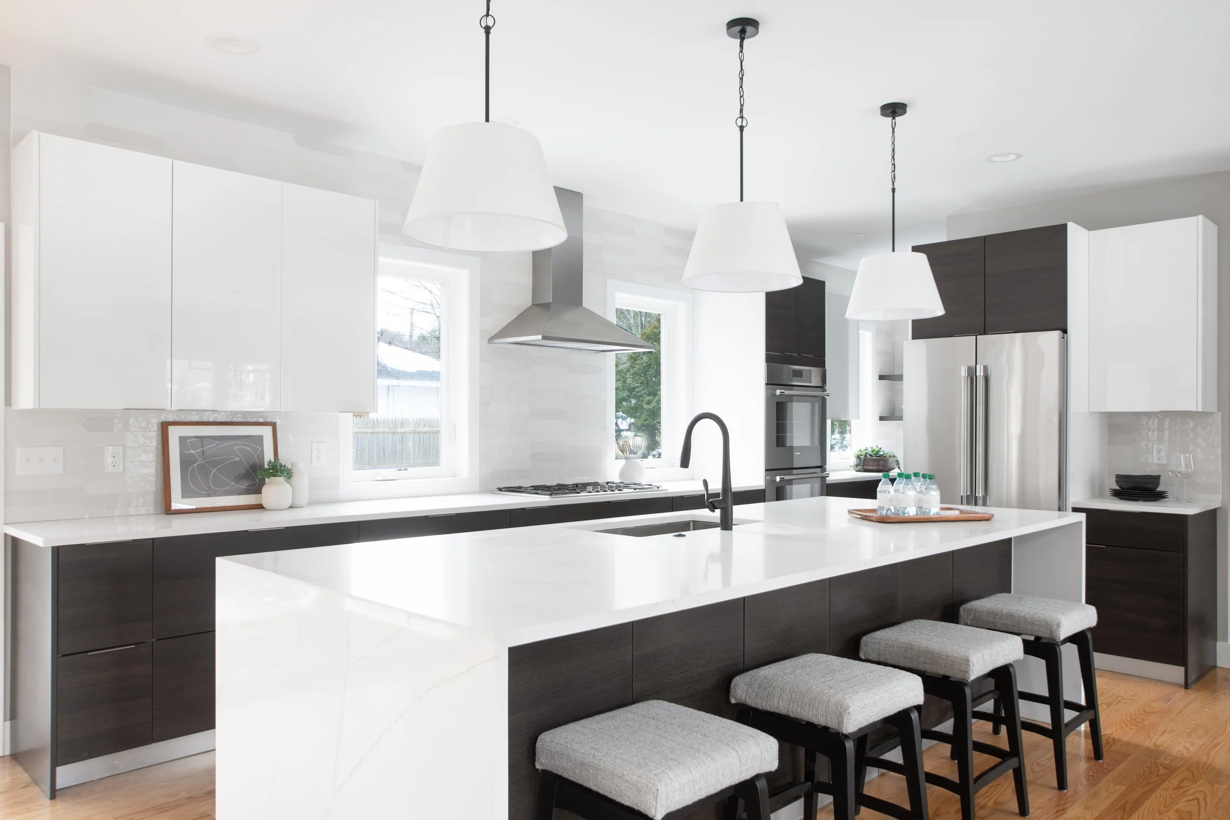Modern kitchen with white countertops, dark cabinetry, stainless steel appliances, and three white pendant lights hanging from the ceiling.