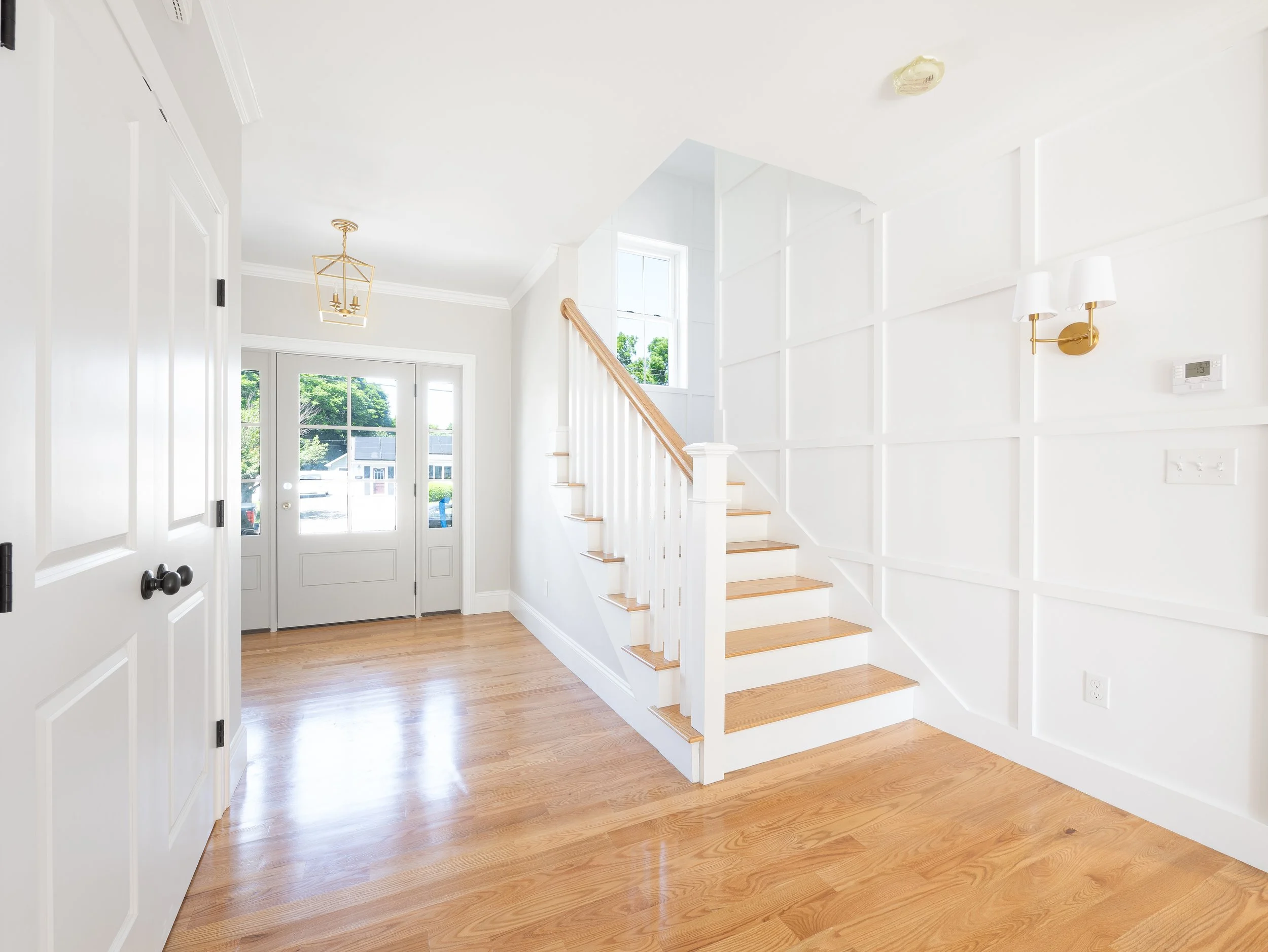 Bright foyer with front door, wooden staircase, hardwood floors, white walls, gold light fixtures, and windows allowing natural light.