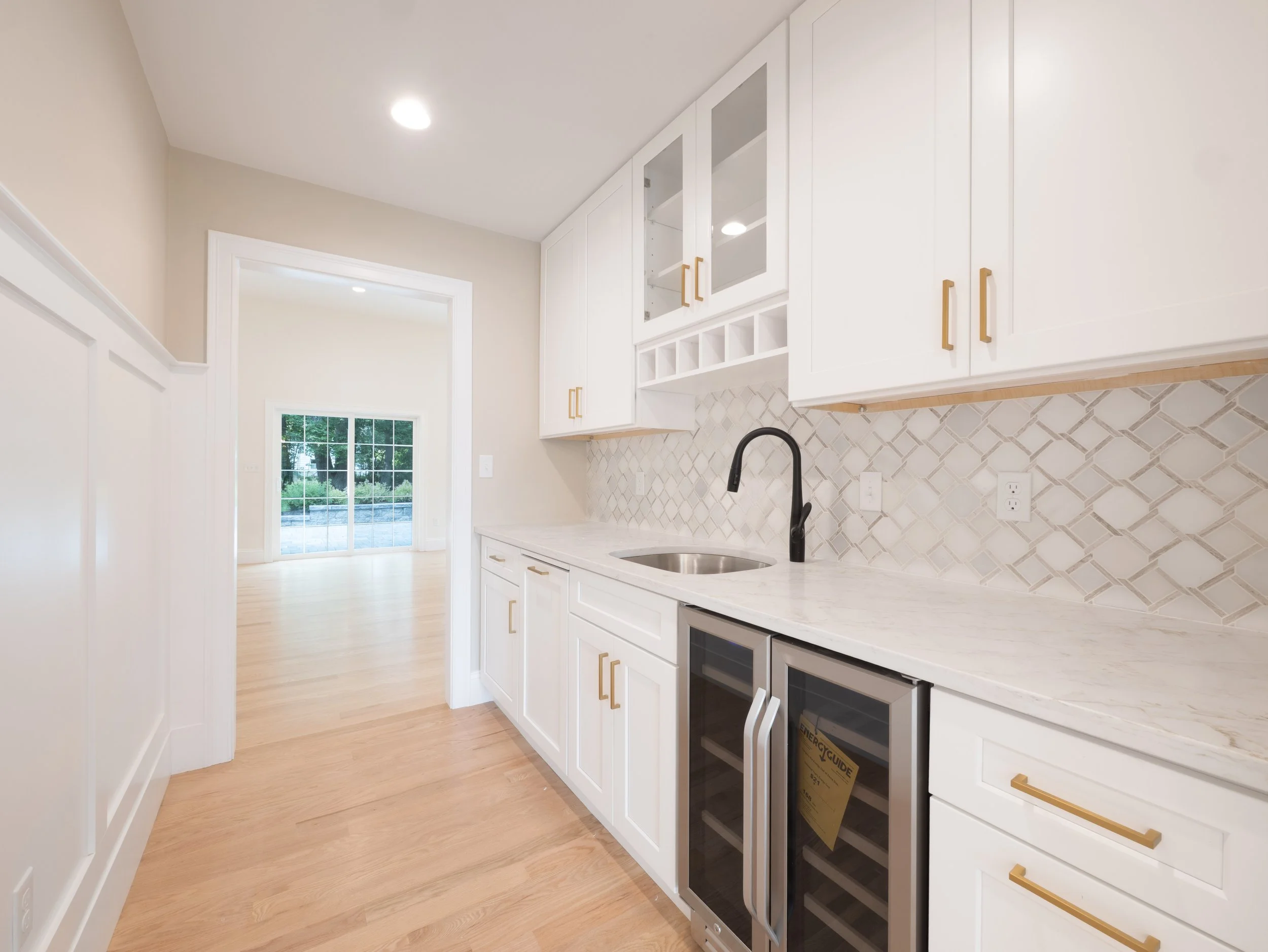 Kitchen with white cabinets, marble countertop, tile backsplash, black faucet, small wine refrigerator, and a view through a sliding glass door to an outdoor area.
