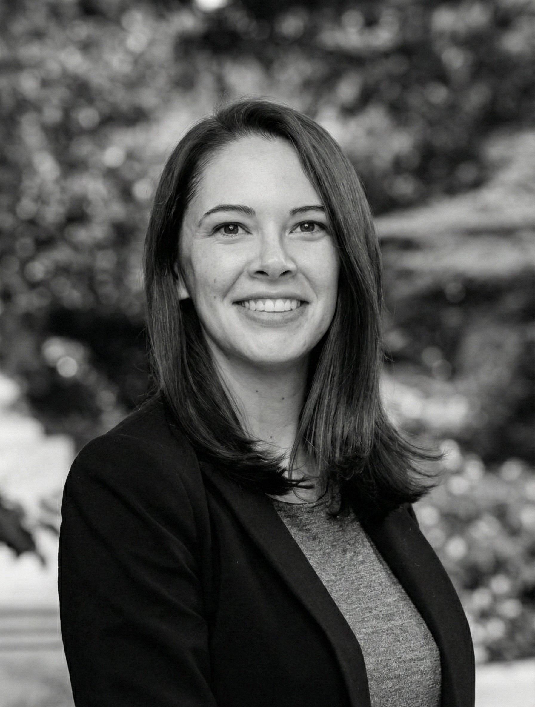 A portrait of a woman with shoulder-length dark hair, smiling, outdoors with blurred trees in the background.