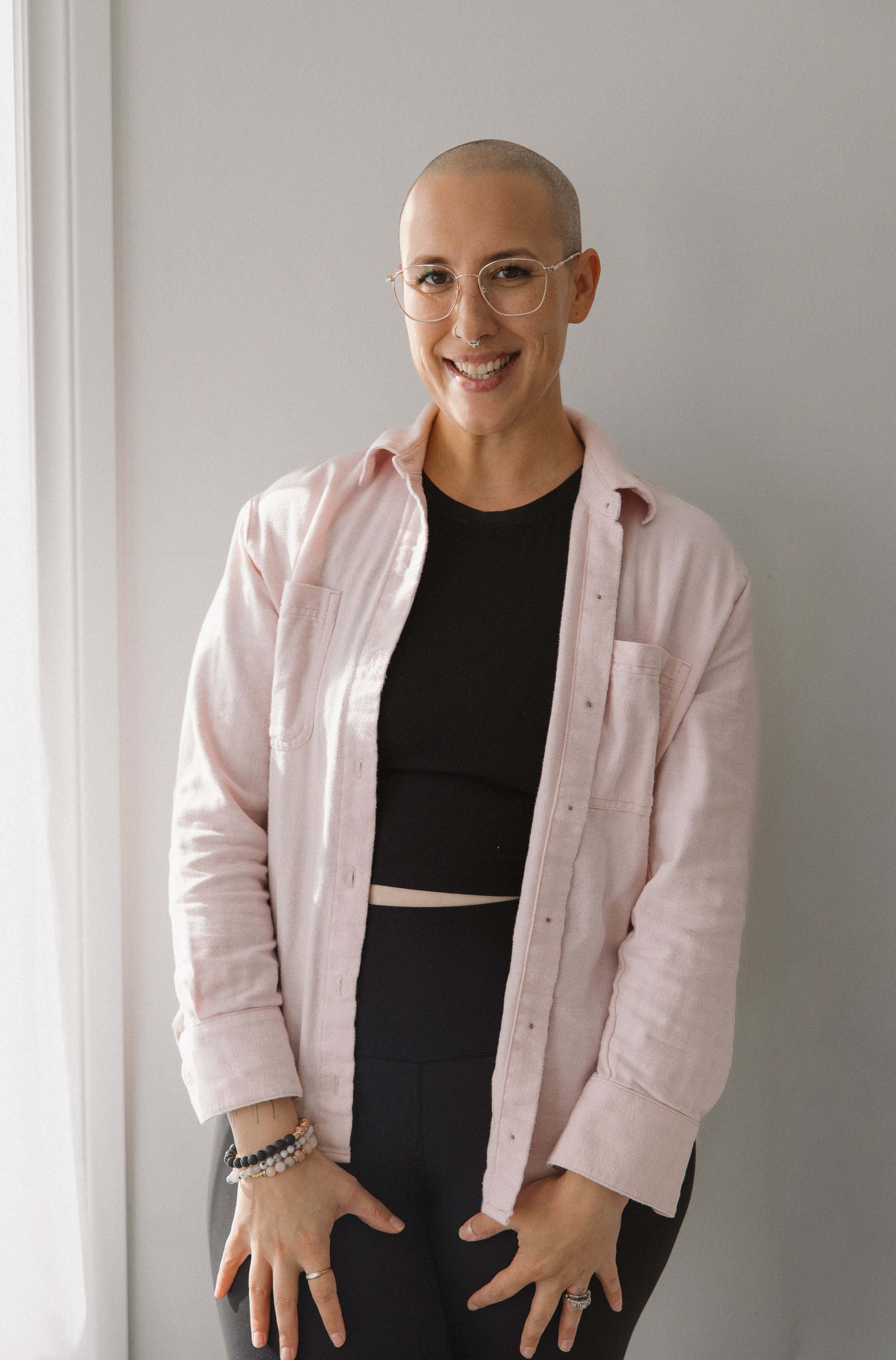 A woman with a shaved head wearing glasses, a black crop top, and a light pink button-up shirt, smiling and standing against a plain light-colored wall.