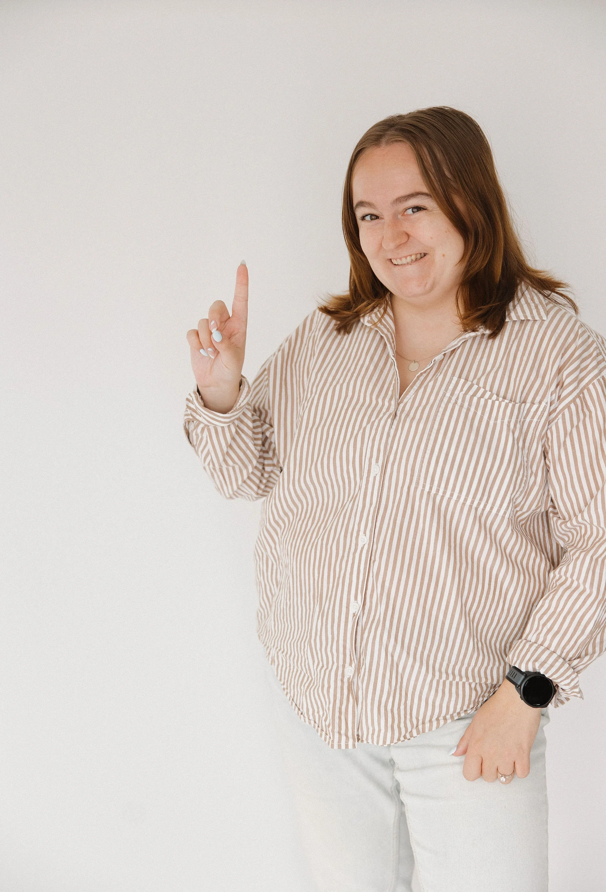 A woman with shoulder-length brown hair, wearing a striped button-up shirt and light-colored pants, smiling, pointing upward with her right index finger.