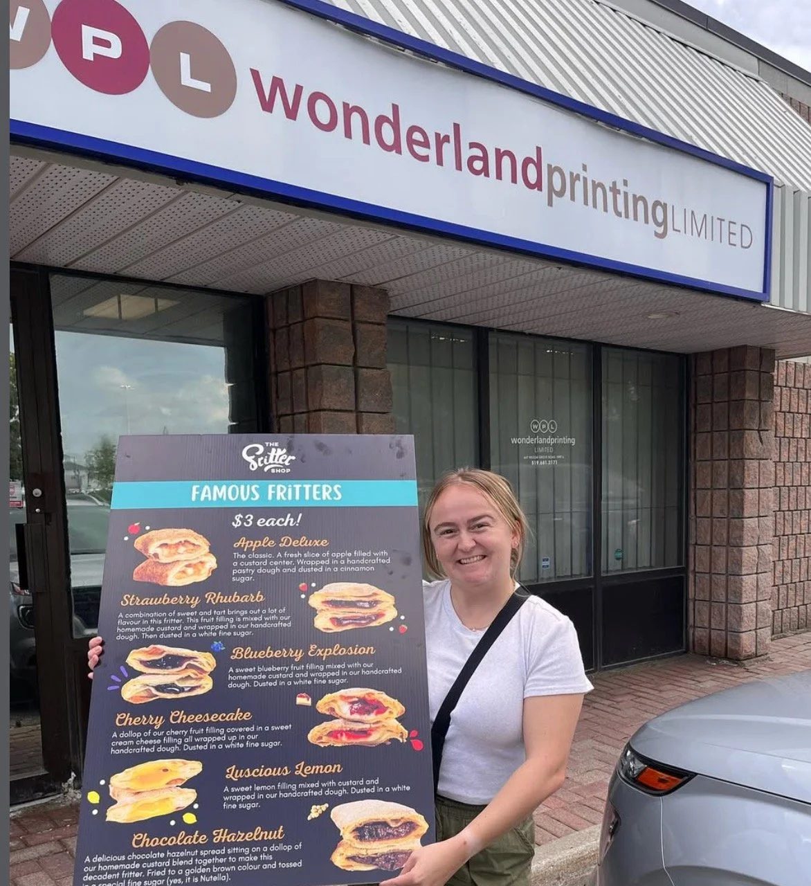 Ashley Rowe holding a client signage piece for The Fritter Shop, standing outside a printing store