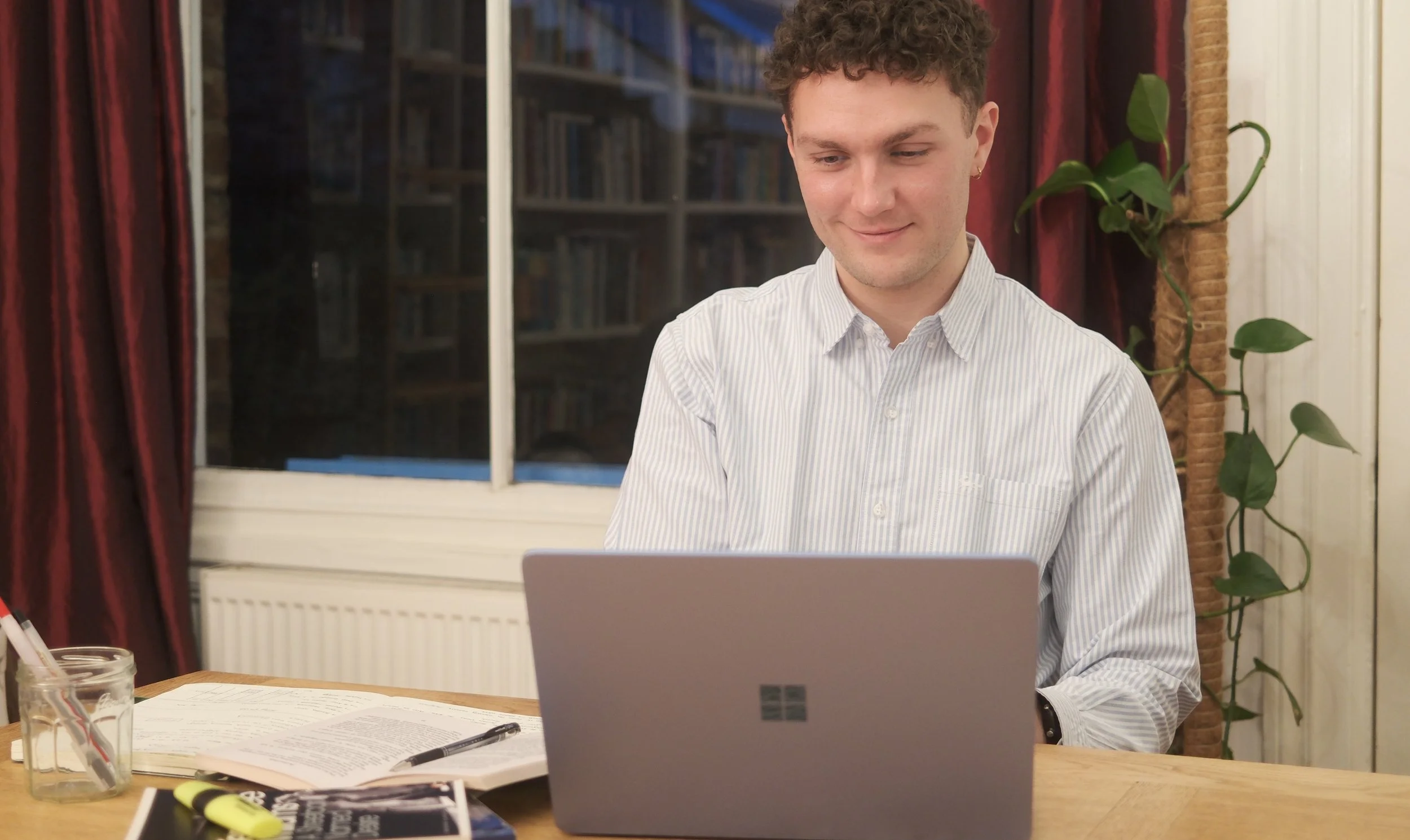 A smiling GCSE and A-Level tutor sitting at a wooden table conducting an English Language / English Literature lesson using his laptop with the exam texts from the syllabus next to him.