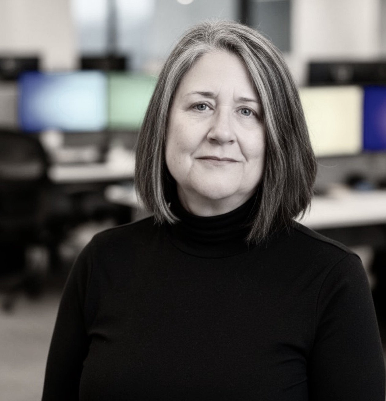 A middle-aged woman with shoulder-length hair wearing a black turtleneck, standing in a modern office with colorful computer screens in the background.