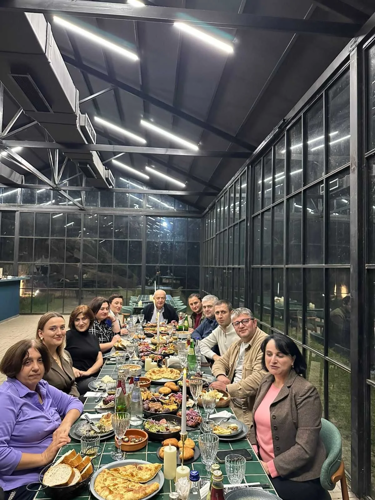 People sitting at a long dinner table with food, drinks, and candles in a greenhouse-like setting.