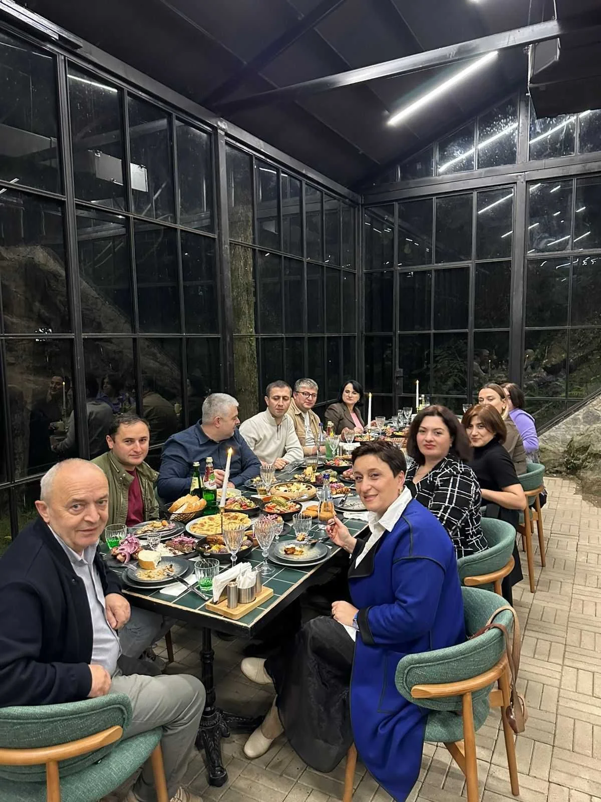 Group of people sitting around a long dining table enjoying a meal in a glass-walled indoor setting.