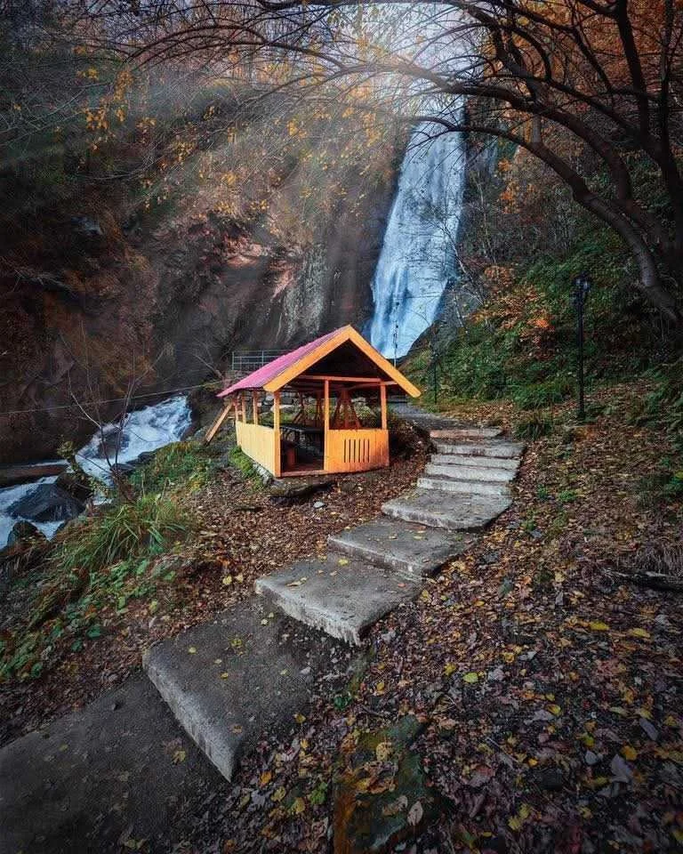 A small pavilion with a red roof and wooden sides built near a waterfall, with stone steps leading up to it and surrounded by fallen autumn leaves.