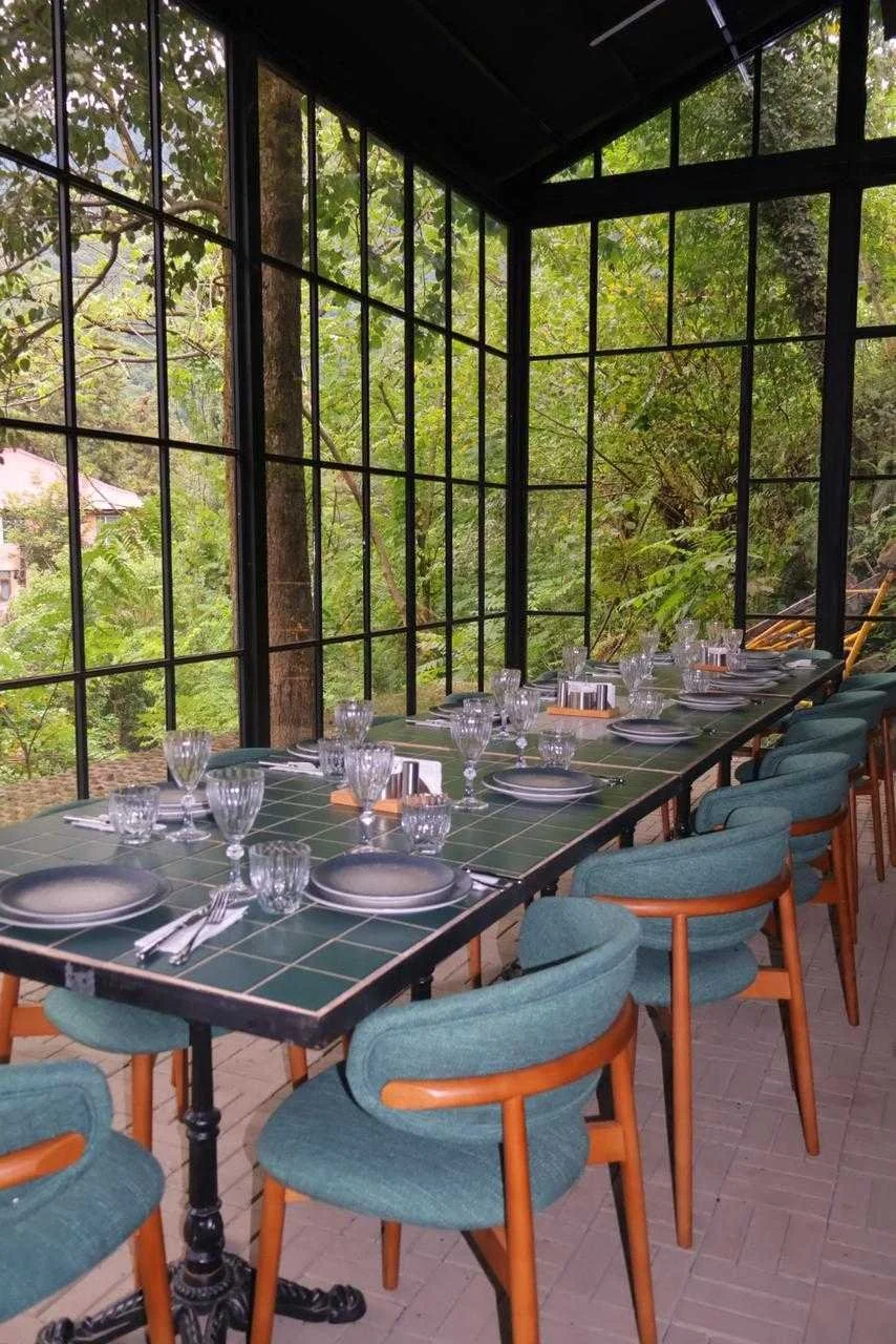 A long dining table set for a meal with plates, glasses, and silverware in a room with floor-to-ceiling windows showing a forest view outdoors.