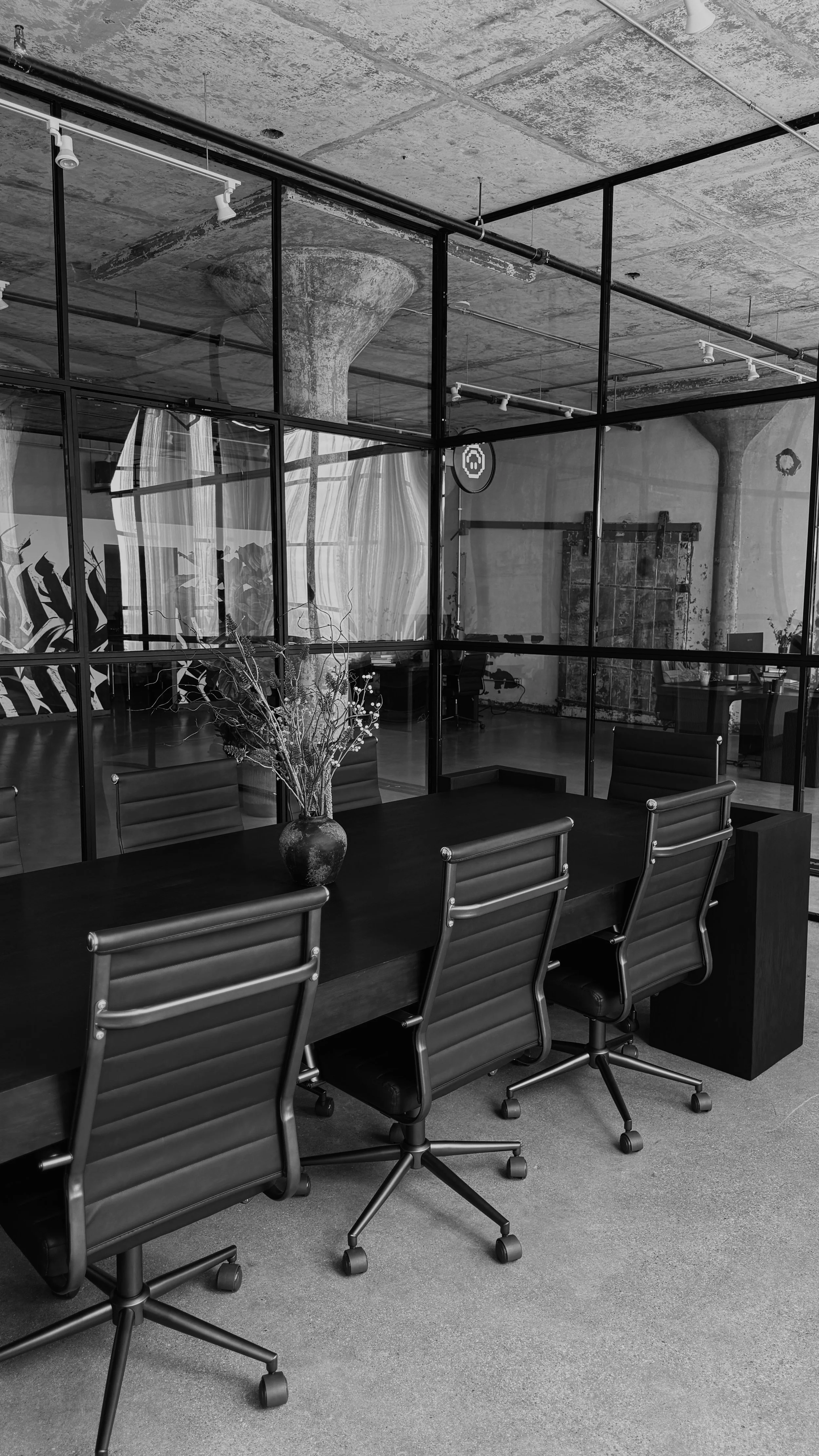 Modern office conference room with a black table, six black leather chairs, and a vase with dried flowers. There is a glass partition behind the table, revealing an industrial-style space with exposed concrete ceiling and support columns.